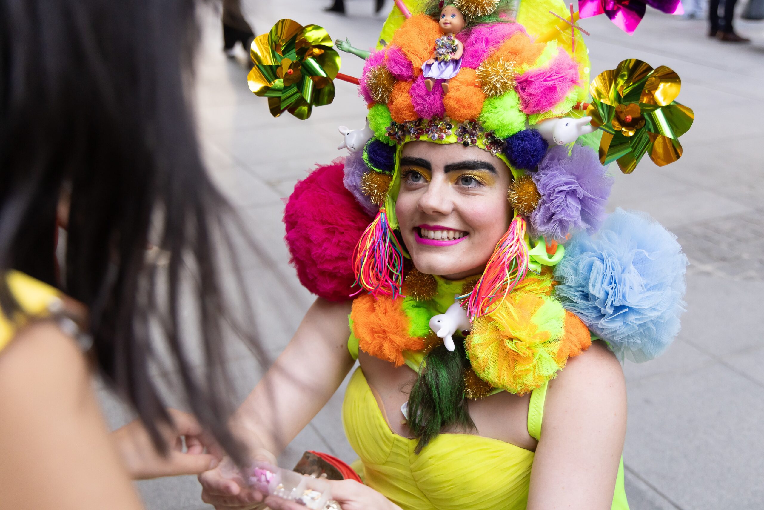 A person in a bright, colorful costume smiles while wearing a vibrant headpiece adorned with fluffy pom-poms, small toys, and shiny decorations. They engage with another person, suggesting a lively, festive atmosphere on a city street.