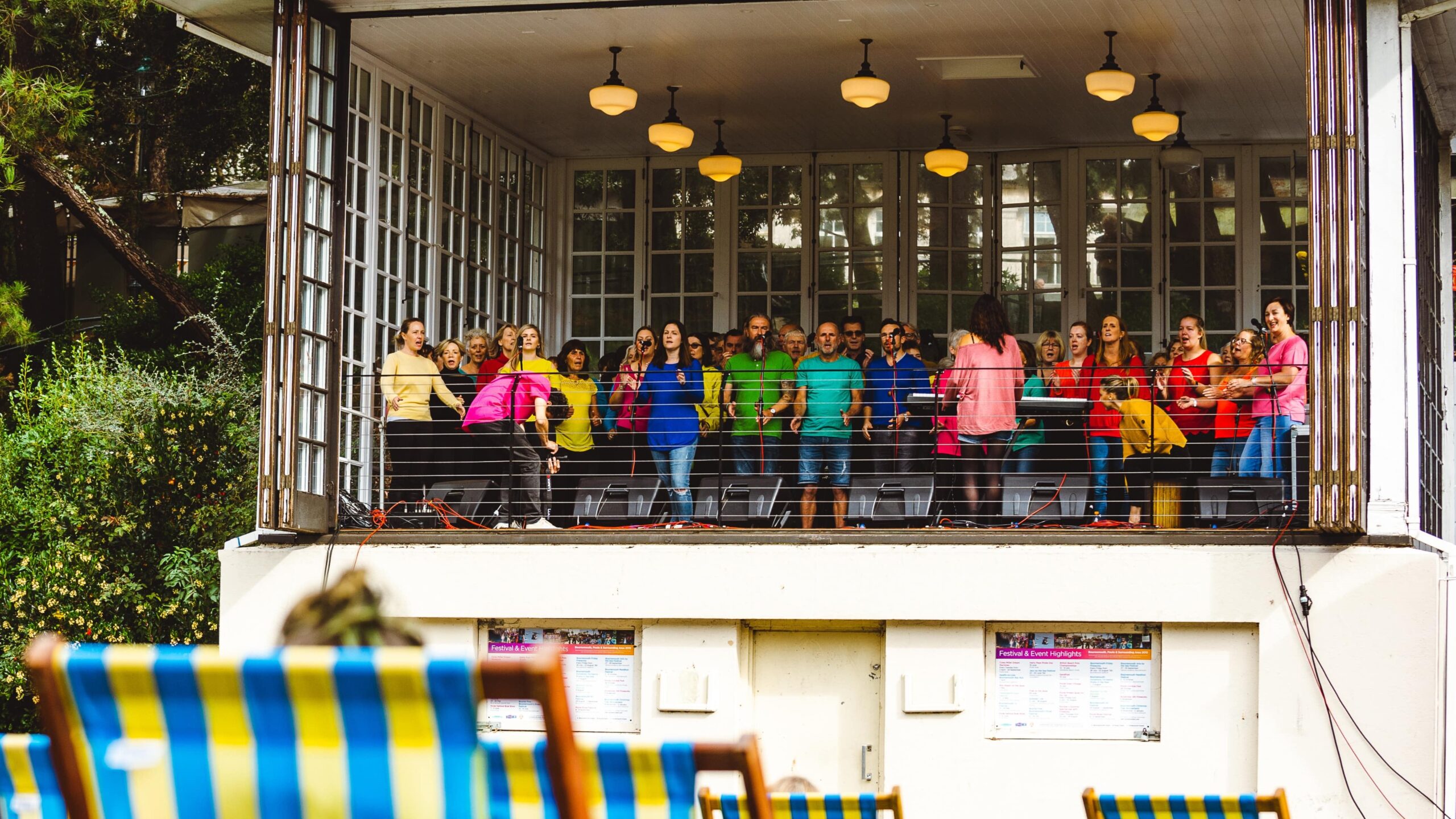 choir of people dressed in brightly colored clothing sings on a raised, enclosed platform while a small audience watches from striped deck chairs on the lawn below.