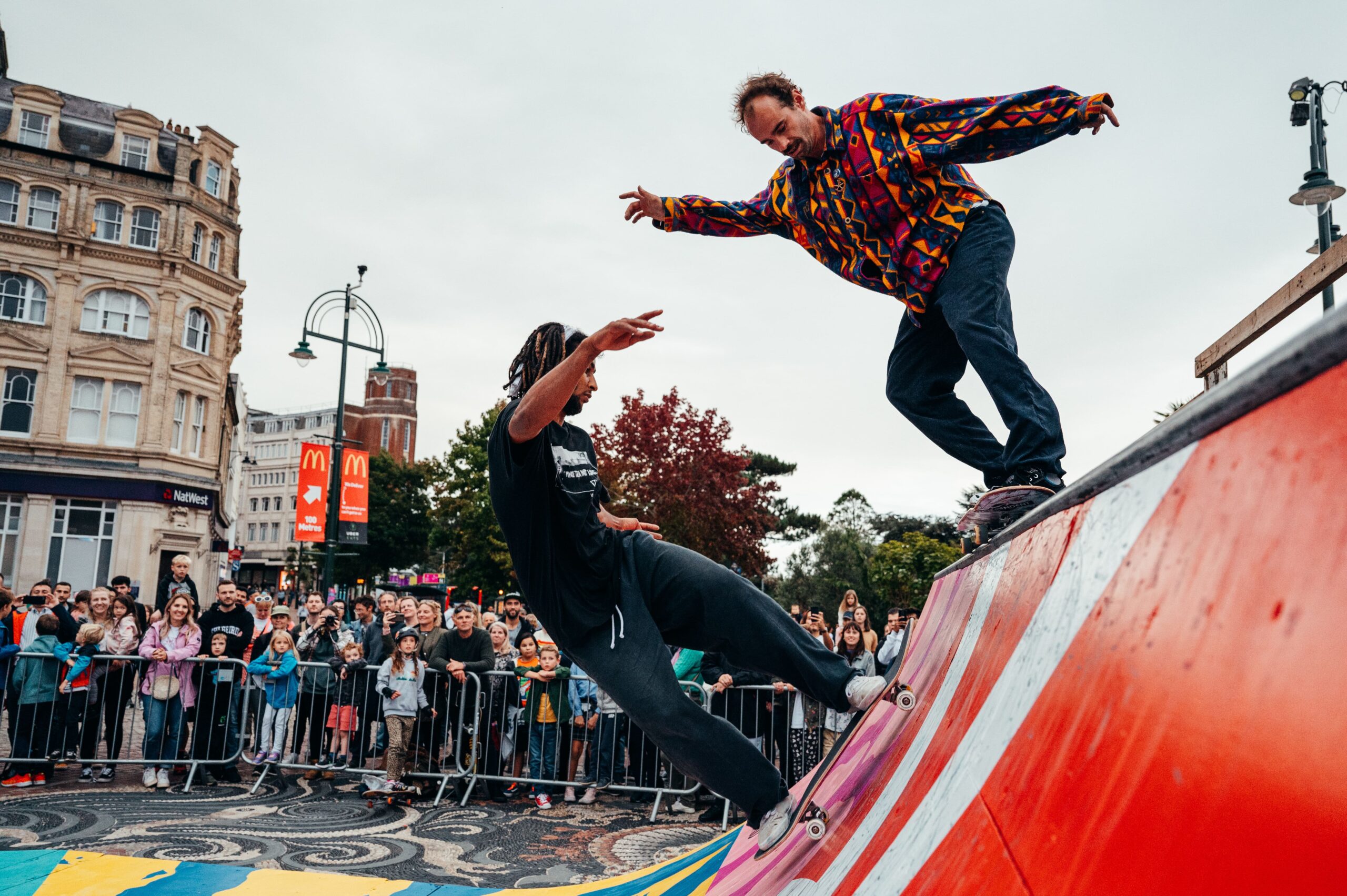 Two skateboarders perform tricks on a colorful ramp as a crowd watches. One skater rides up the ramp in black clothing, while the other balances on the edge wearing a vibrant shirt. The scene is set in an urban area with historic buildings and a McDonald's sign in the background.