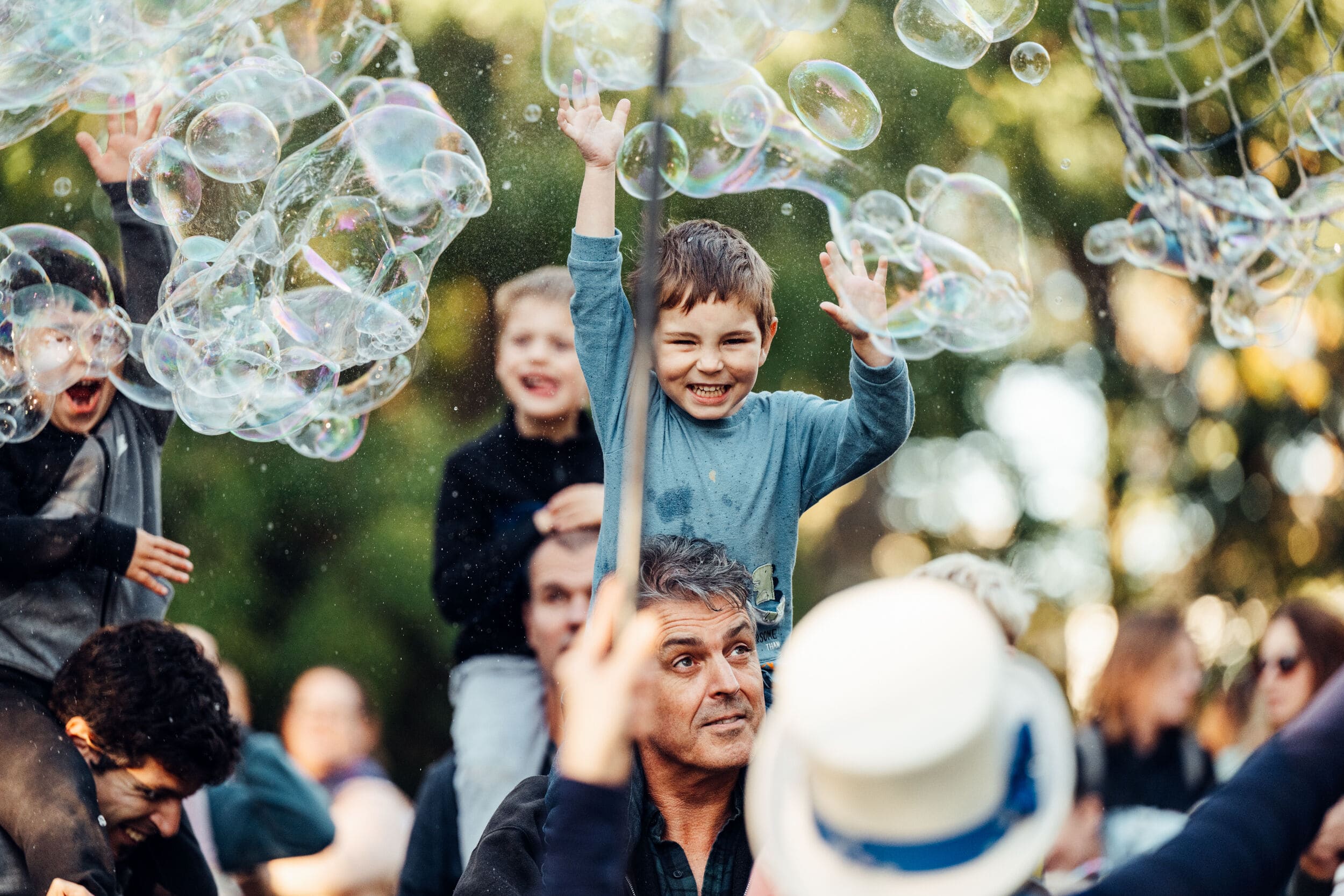 A joyful child sits on someone's shoulders, reaching up to pop large, iridescent bubbles floating in the air. Other children in the background also eagerly reach for the bubbles, their faces lit with excitement. The scene takes place outdoors, with soft-focus greenery and a lively crowd in the background.