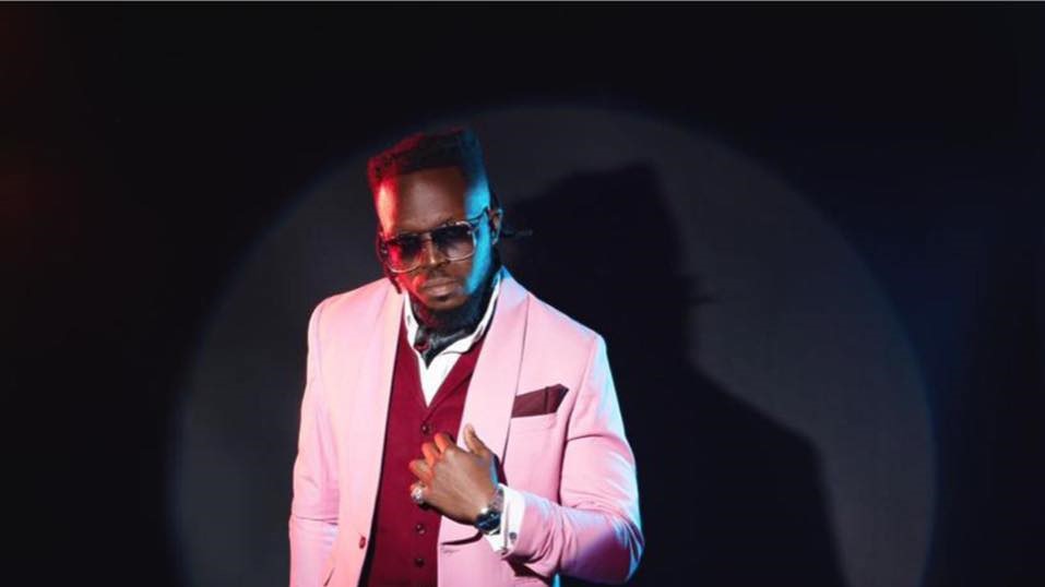 A man stands confidently against a dark backdrop, illuminated by spotlight, wearing a stylish pink suit with a red vest and a white shirt. He accessorizes with sunglasses, rings, a watch, and a pocket square