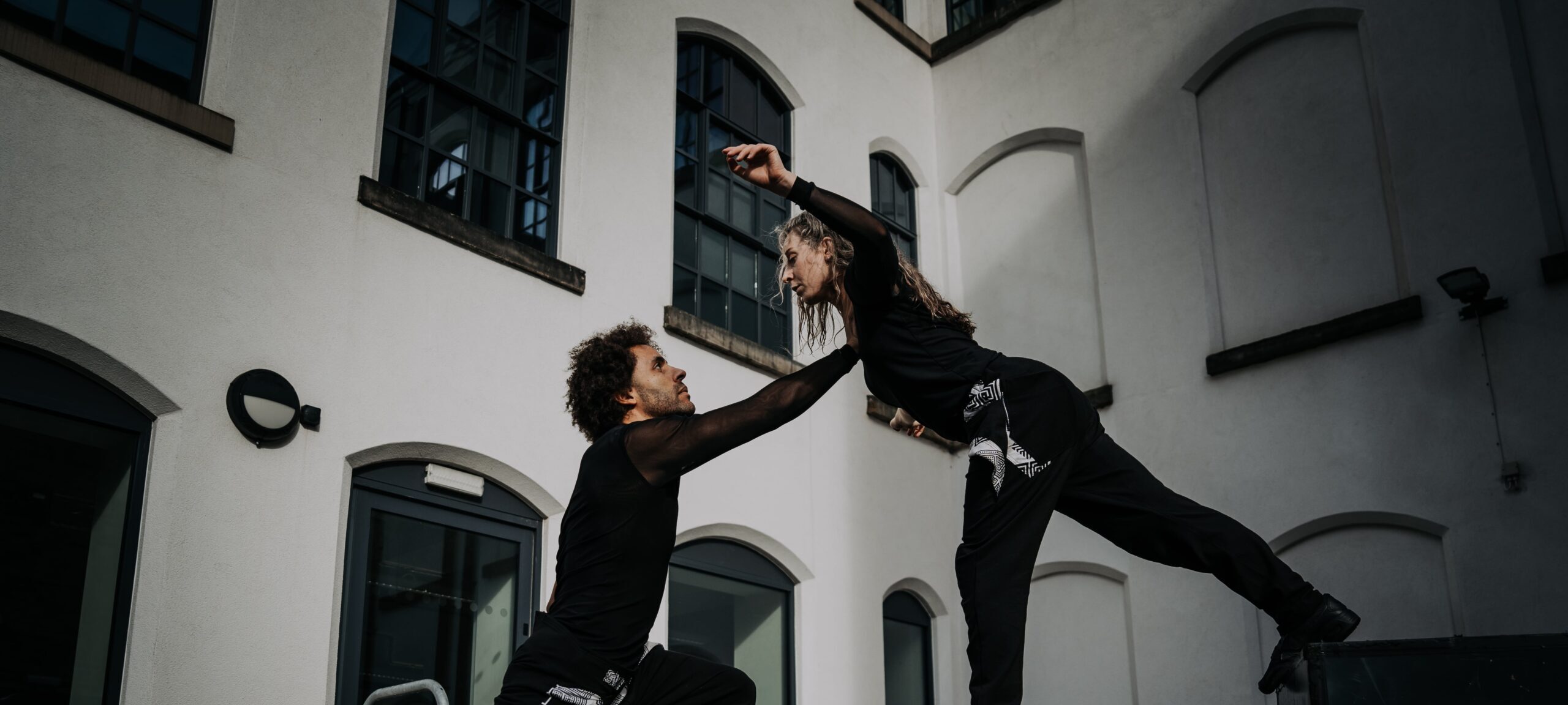 Two dancers performing an elegant move outdoors, with one dancer supporting the other mid-air. Both are dressed in black, and their expressive posture is set against the backdrop of a modern building with large window