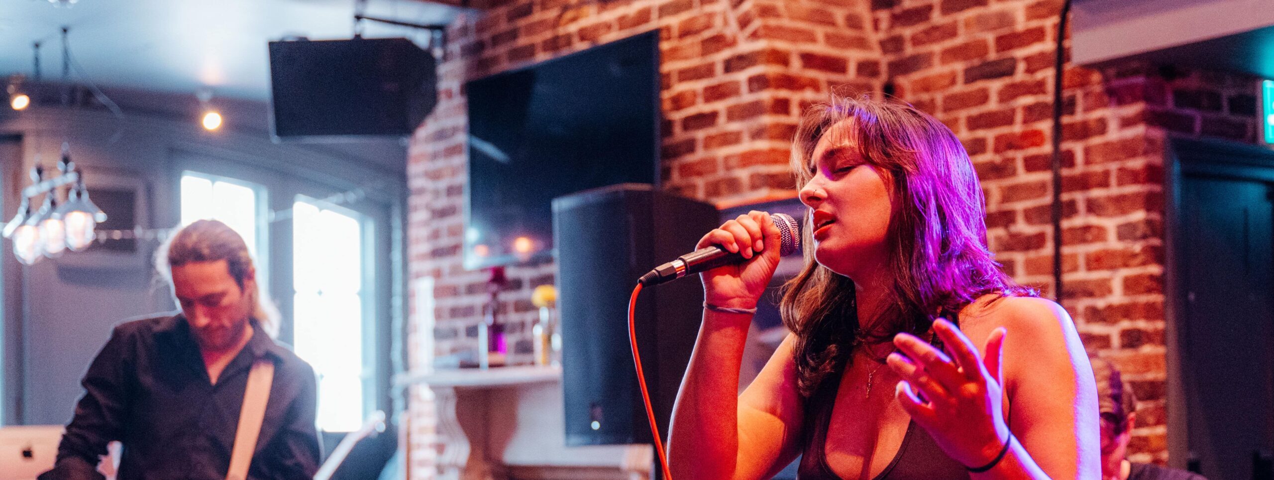 A woman stands at a bar, singing into a microphone, captivating the crowd with her performance in a vibrant atmosphere.