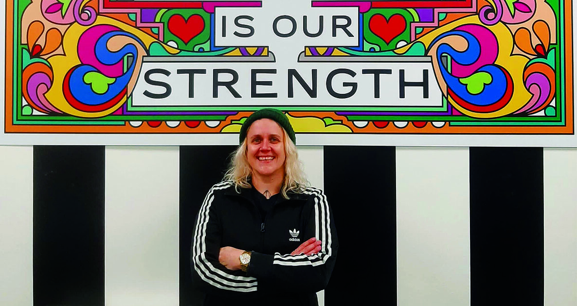 A person with long blond hair, wearing a black Adidas jacket and a beanie, stands smiling with arms crossed in front of a colorful mural that reads, 