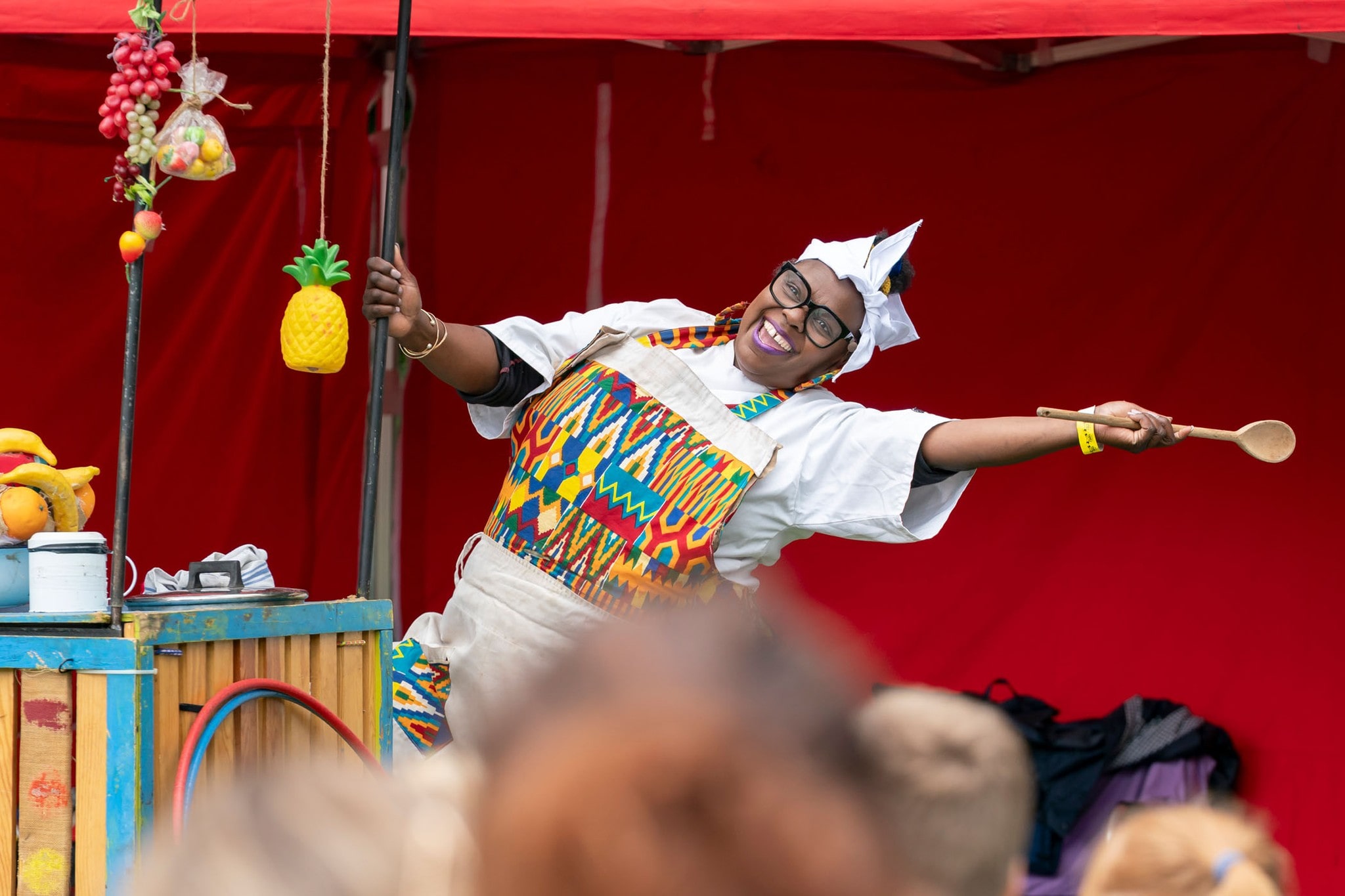 A joyful performer in a colorful, patterned apron and white headscarf is smiling widely while holding a wooden spoon and a pole with a hanging plastic pineapple. They are posing energetically in front of a red canopy, with artificial fruits and a hula hoop visible on a wooden structure nearby, suggesting a lively market or festival scene.