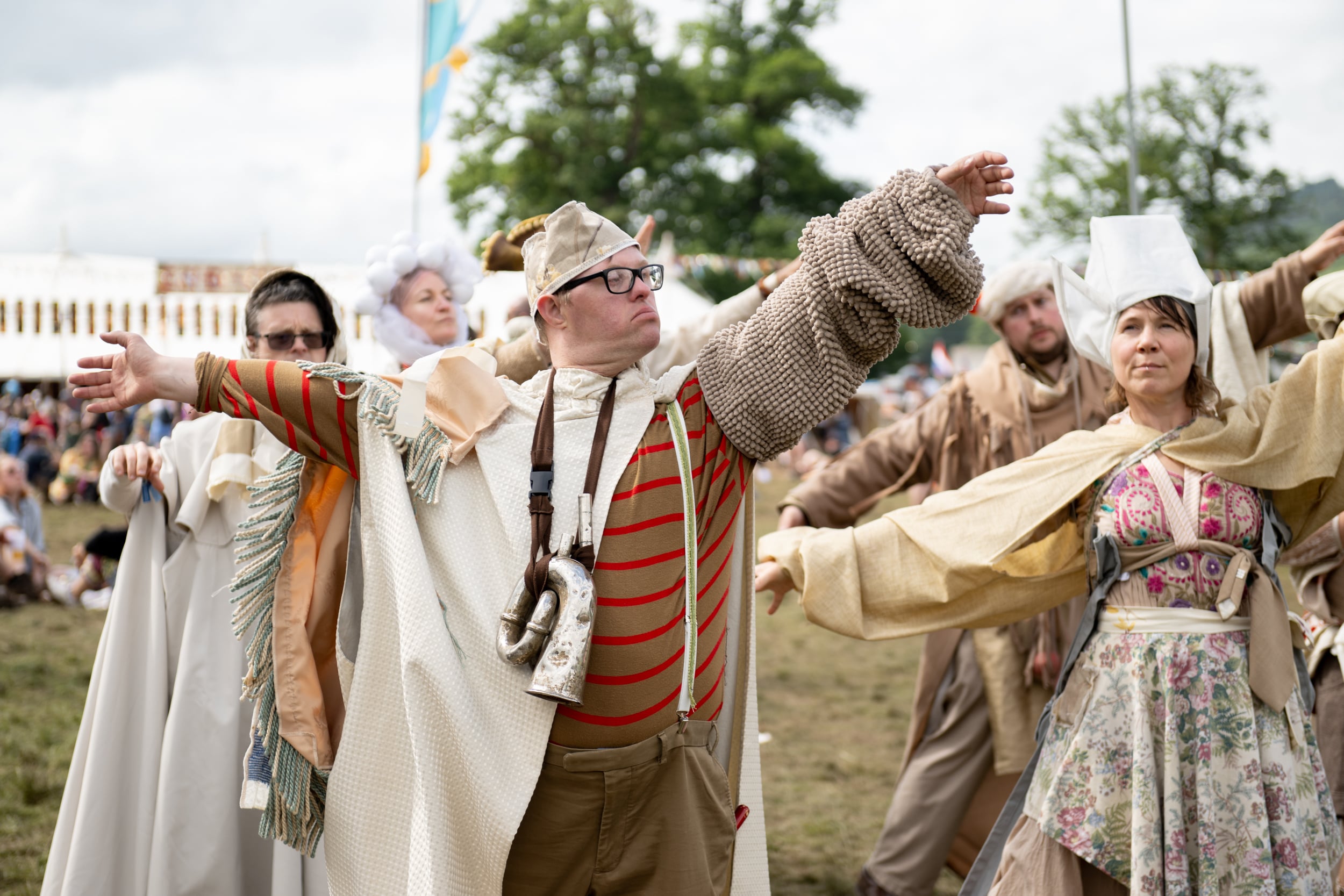 A group of performers in historical-inspired costumes stand with their arms outstretched in a dramatic pose at an outdoor festival, with tents and spectators in the background.