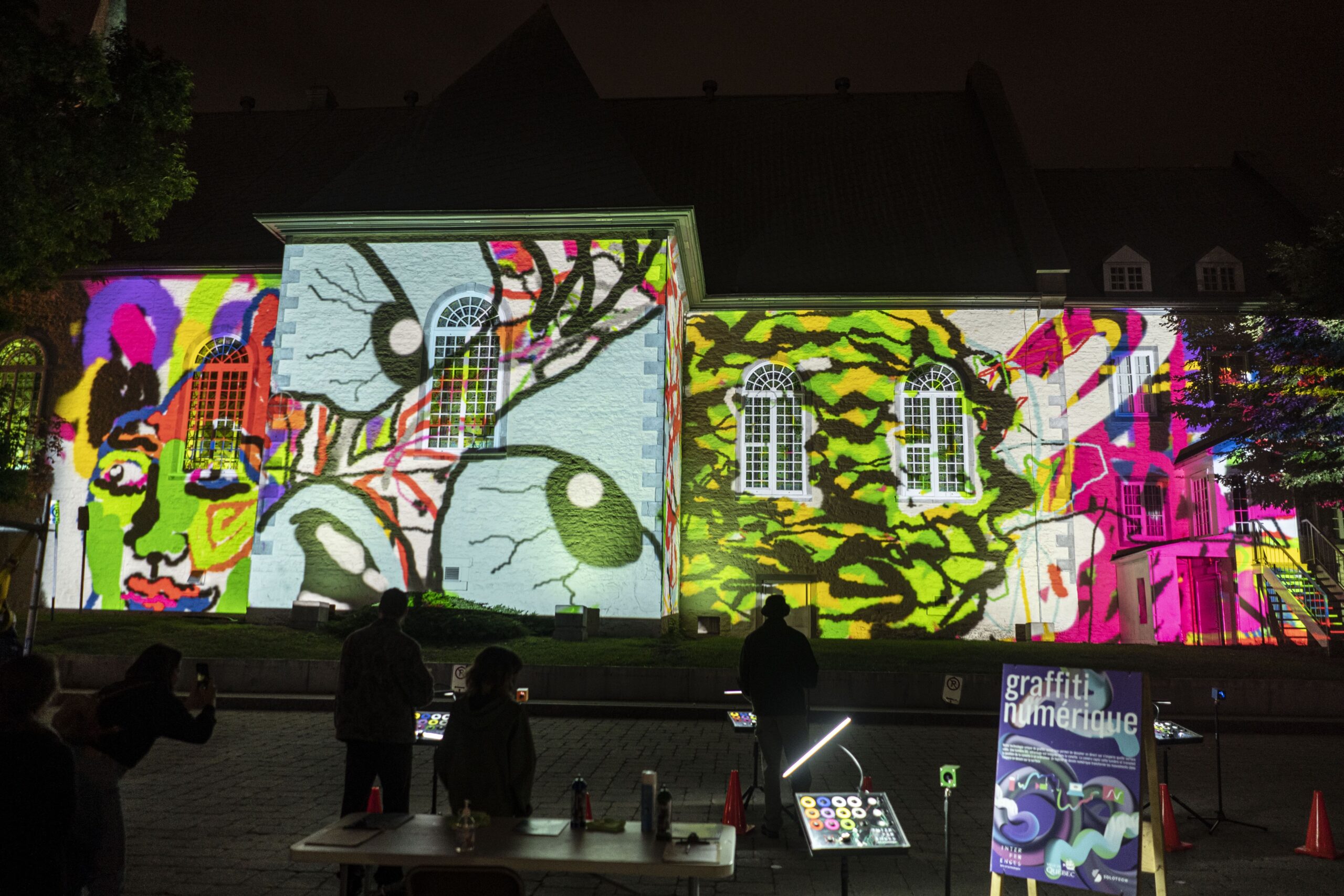 a vibrant light projection on the exterior of a building at night. The facade is illuminated with colorful, abstract graffiti-like patterns, including faces and organic shapes. People are seen observing and possibly interacting with the digital artwork, adding to the dynamic, immersive atmosphere.