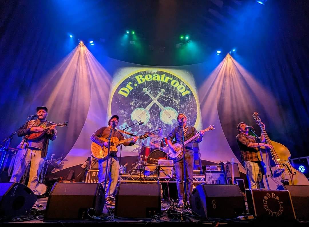 A band performs on stage under colorful lighting, with a backdrop displaying the name 