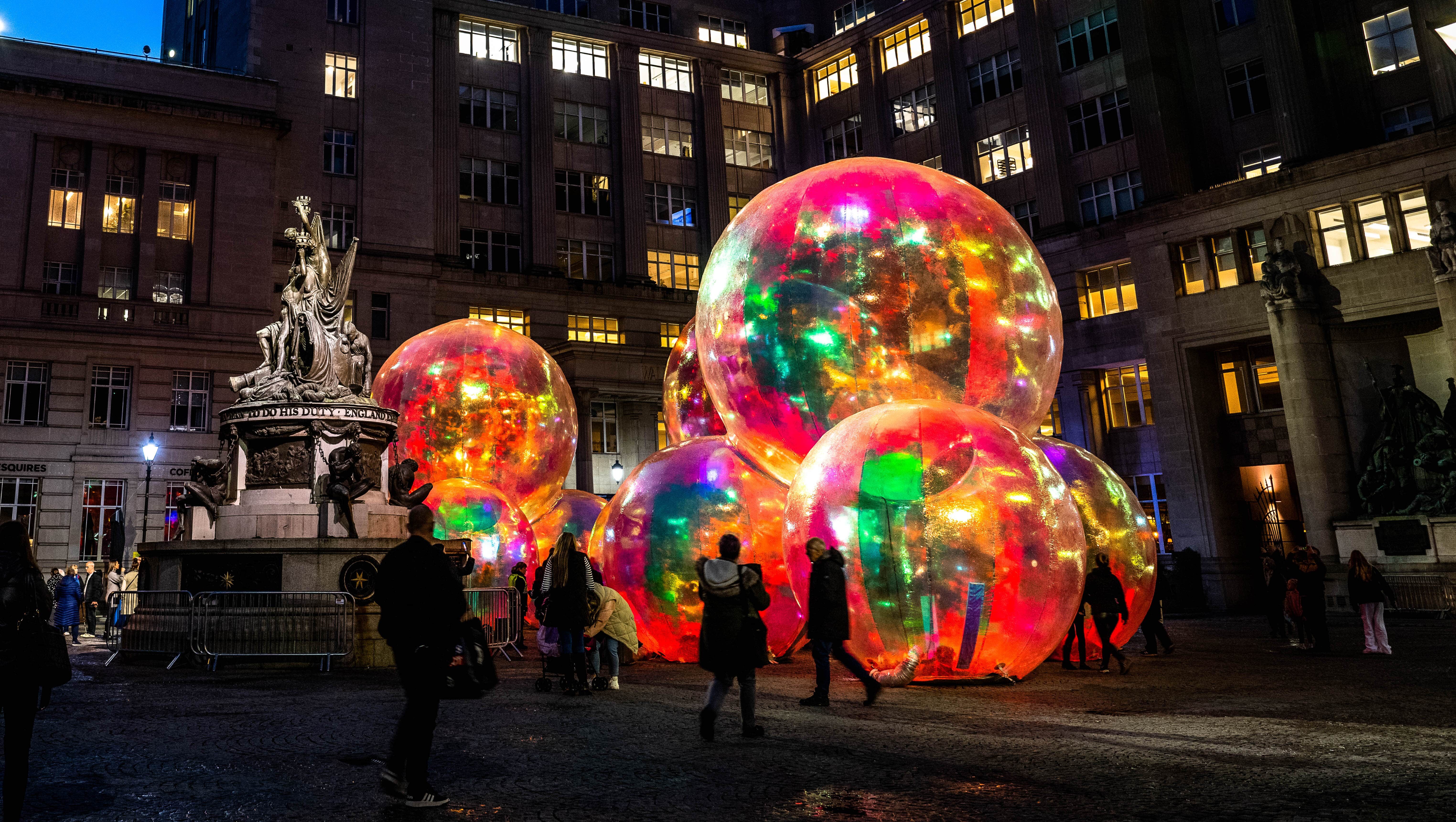 A nighttime scene featuring large, colorful, glowing orbs in an urban square, surrounded by tall buildings and a historic statue, with people admiring the installation.