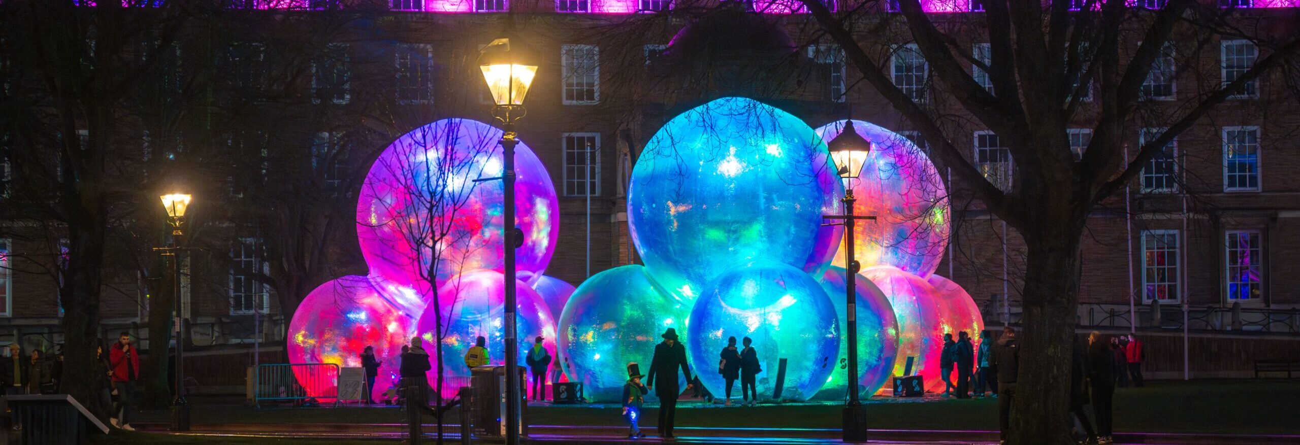 An outdoor nighttime scene featuring a group of large, transparent, iridescent inflatable bubbles illuminated with blue, green, and pink light, creating a glowing effect.