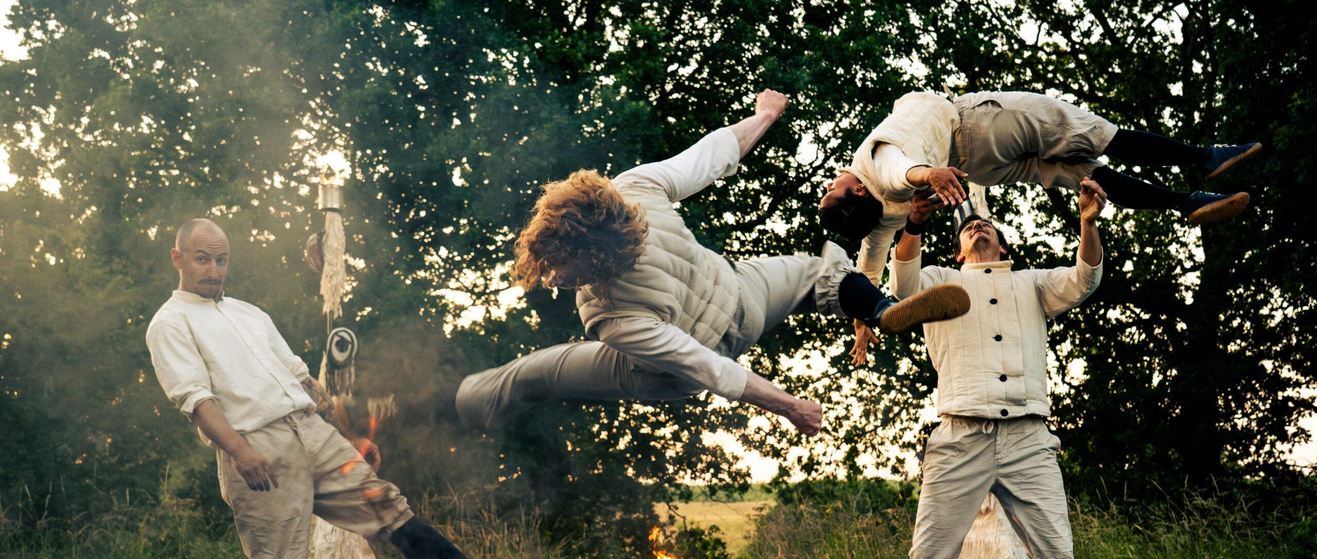 Four performers dressed in white and beige clothing are engaged in a dynamic acrobatic routine outdoors. Two performers are airborne, mid-flip, while others interact on the ground. Flames and smoke enhance the dramatic effect of the scene.