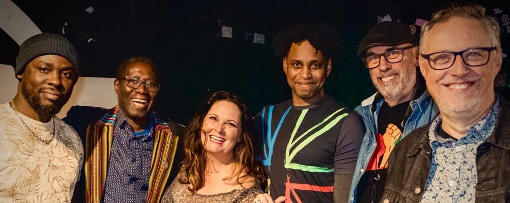 A group of six people stands together, smiling for a group photo. They are dressed in a variety of styles, with the backdrop being a dimly lit room that suggests a casual, friendly gathering.