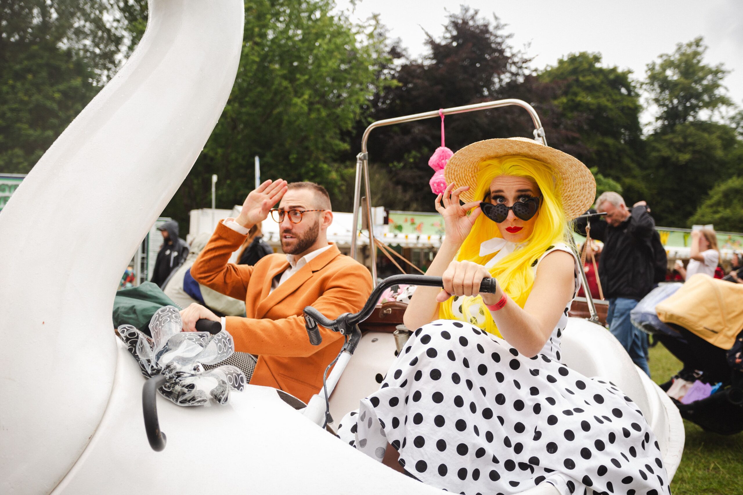 A man in an orange suit and glasses and a woman with bright yellow hair, a straw hat, and a polka-dot dress are riding in a swan-shaped vehicle.