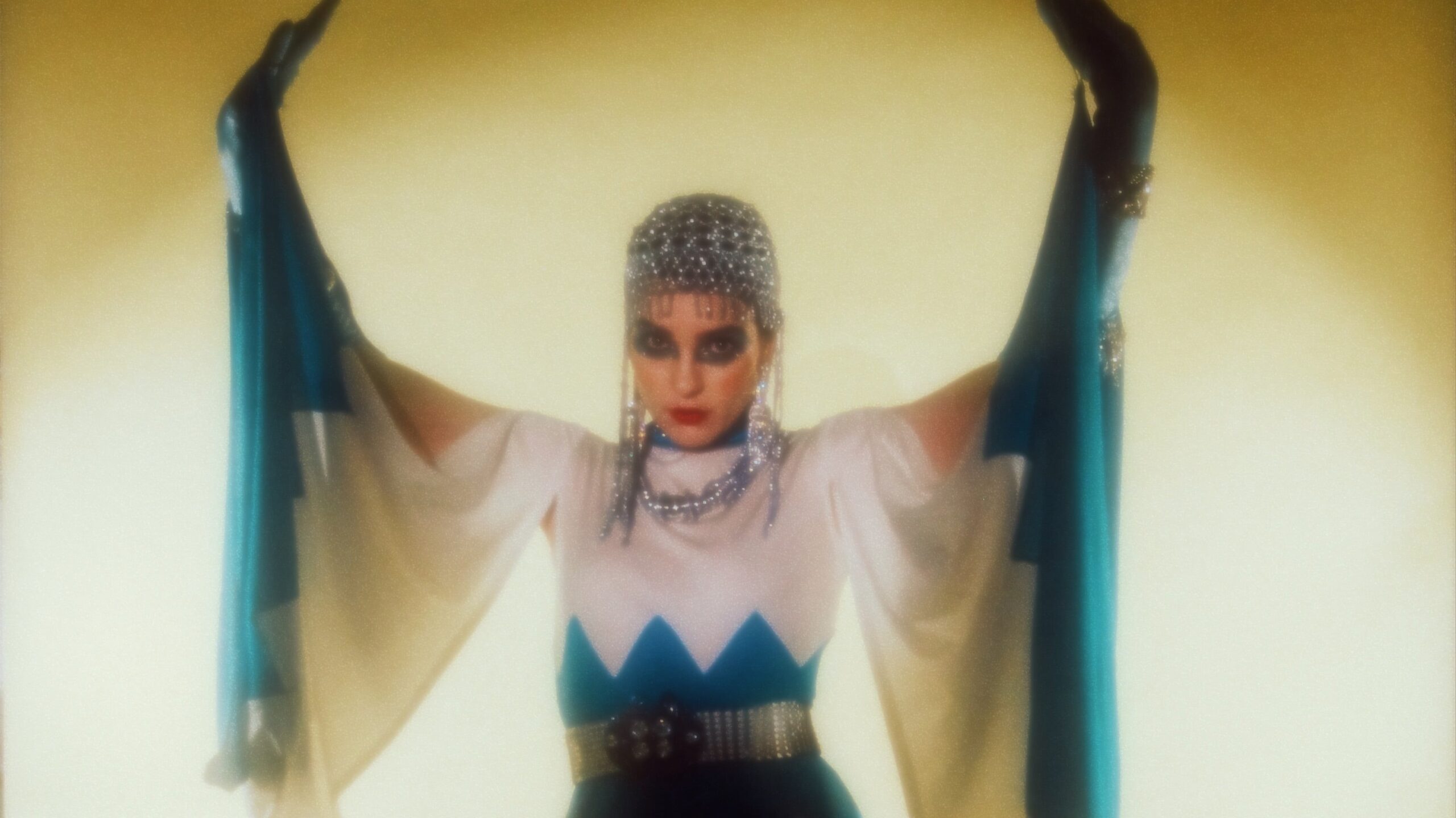 A person poses dramatically against a yellow background, wearing a costume with flowing sleeves, a beaded headpiece, and a wide belt.