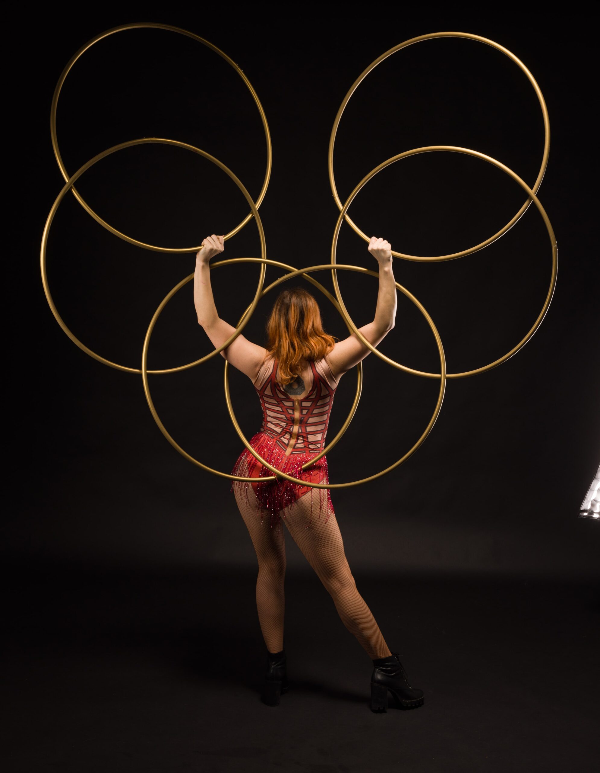 A woman with long red hair stands with her back to the camera, holding up multiple large golden hoops. She is wearing a form-fitting, red, sparkly costume