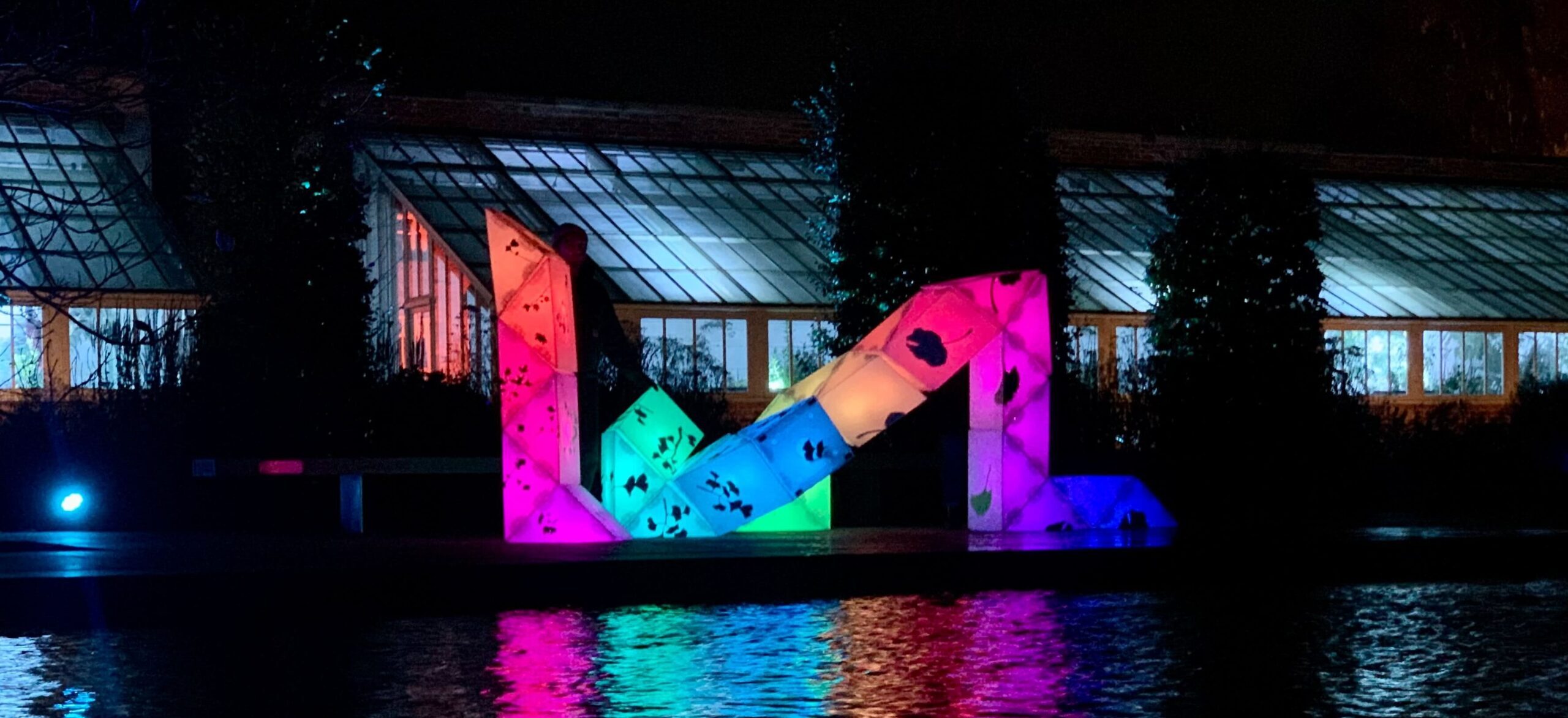 A nighttime scene featuring a vibrant light installation near a water body. The installation is illuminated with bright colors like pink, blue, and green, reflecting beautifully on the water surface.