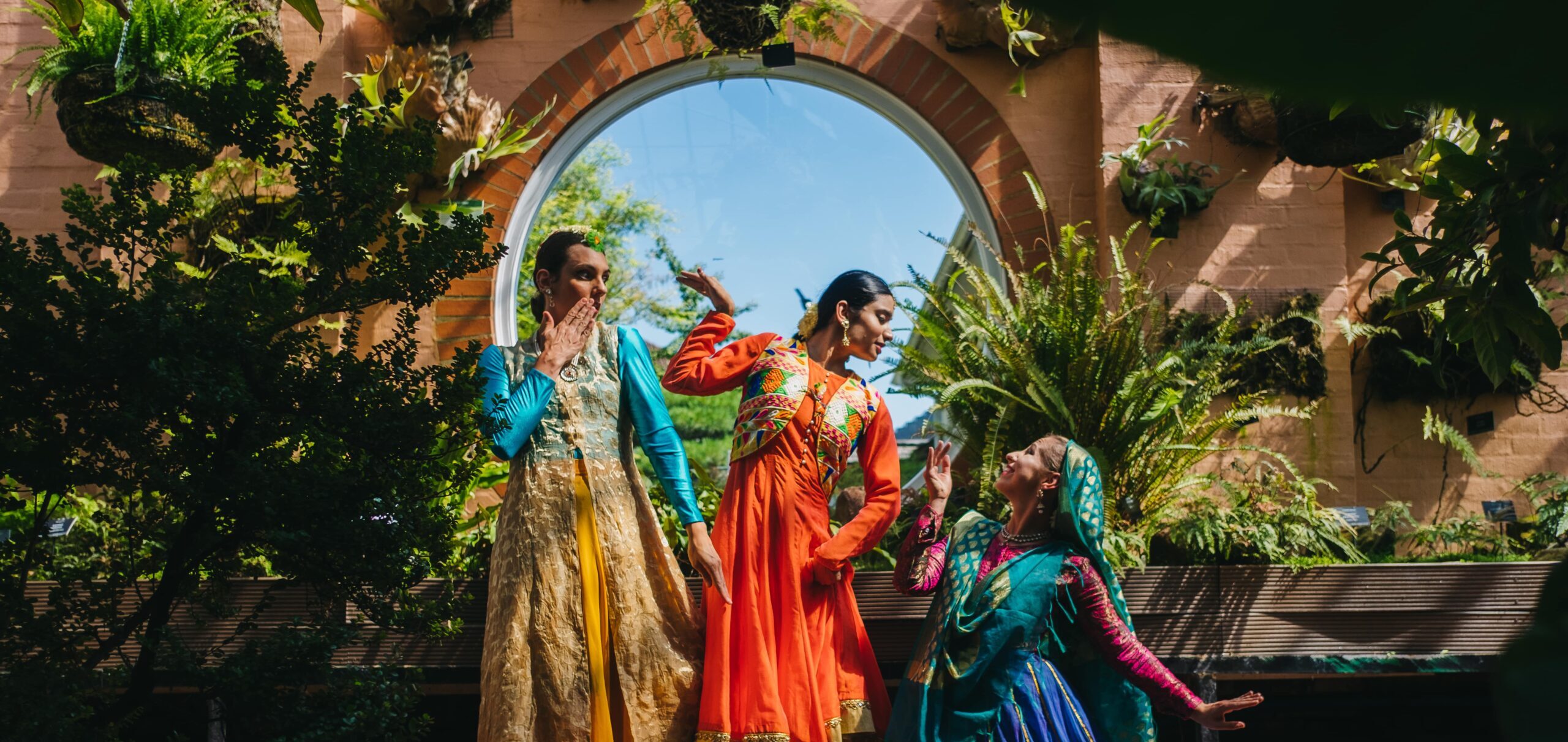 Three dancers in colorful, traditional Mughal-inspired costumes perform indoors, surrounded by lush greenery. The setting includes a large, round window that brings natural light into the space