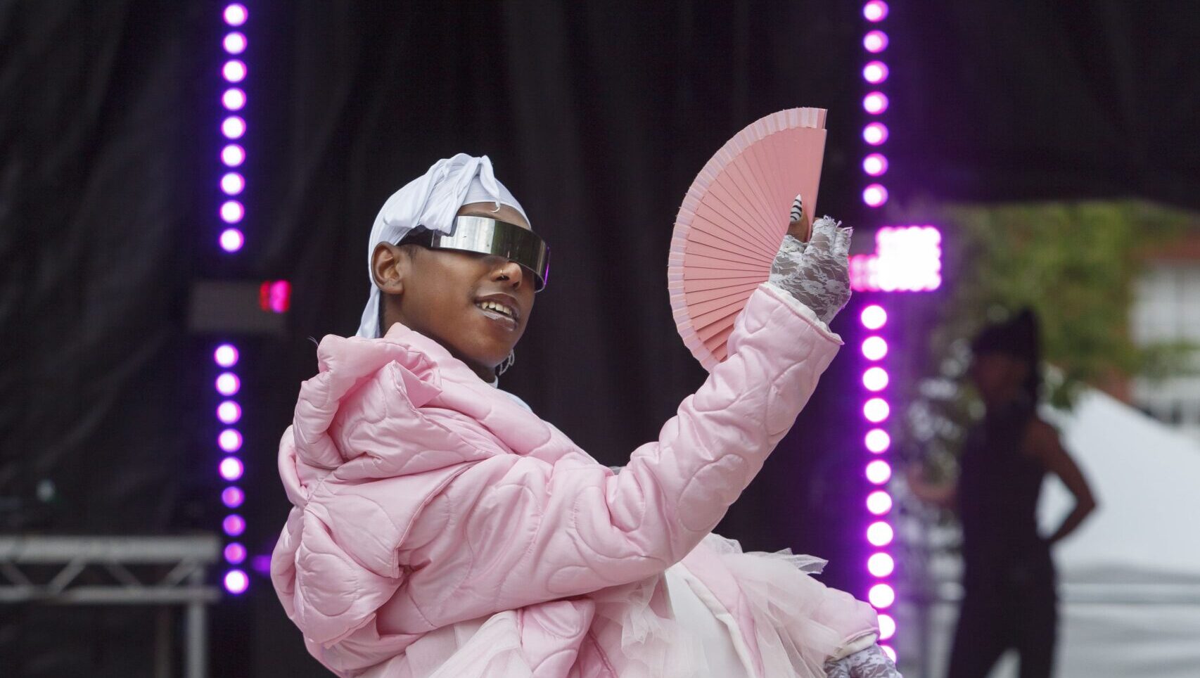 A person wearing a pink dress and sunglasses stands confidently on a stage, exuding elegance and style.