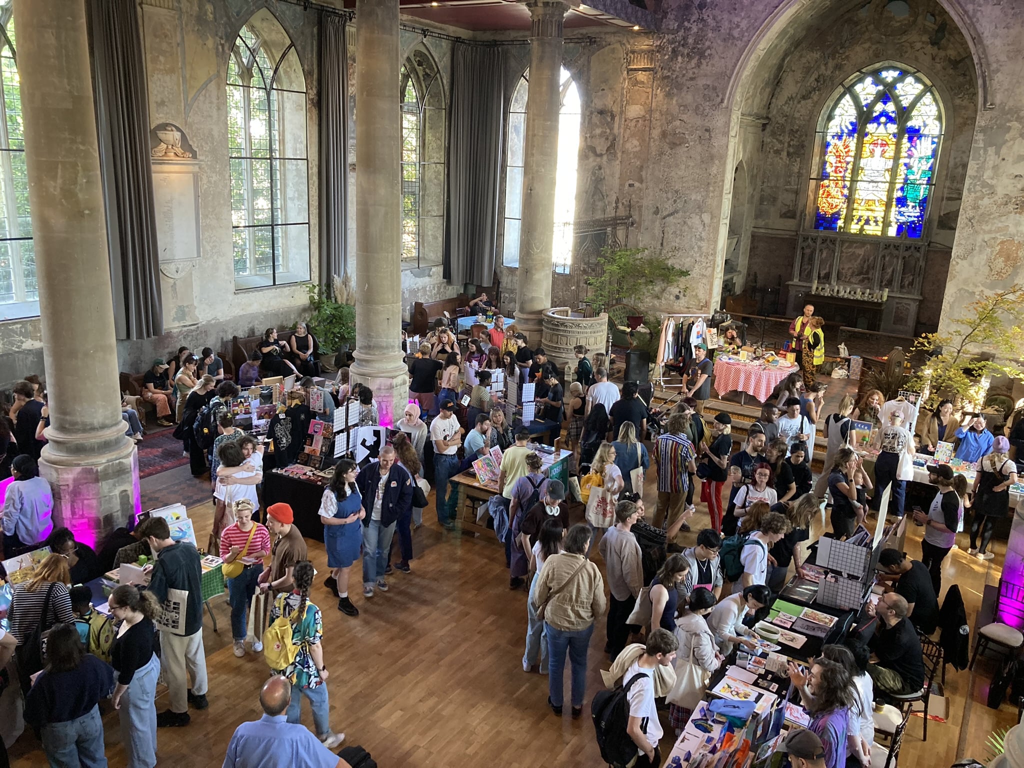 An overhead view of a bustling indoor market held in a historic building with tall columns and large arched windows. The space is filled with people browsing various stalls, creating a lively and vibrant atmosphere.