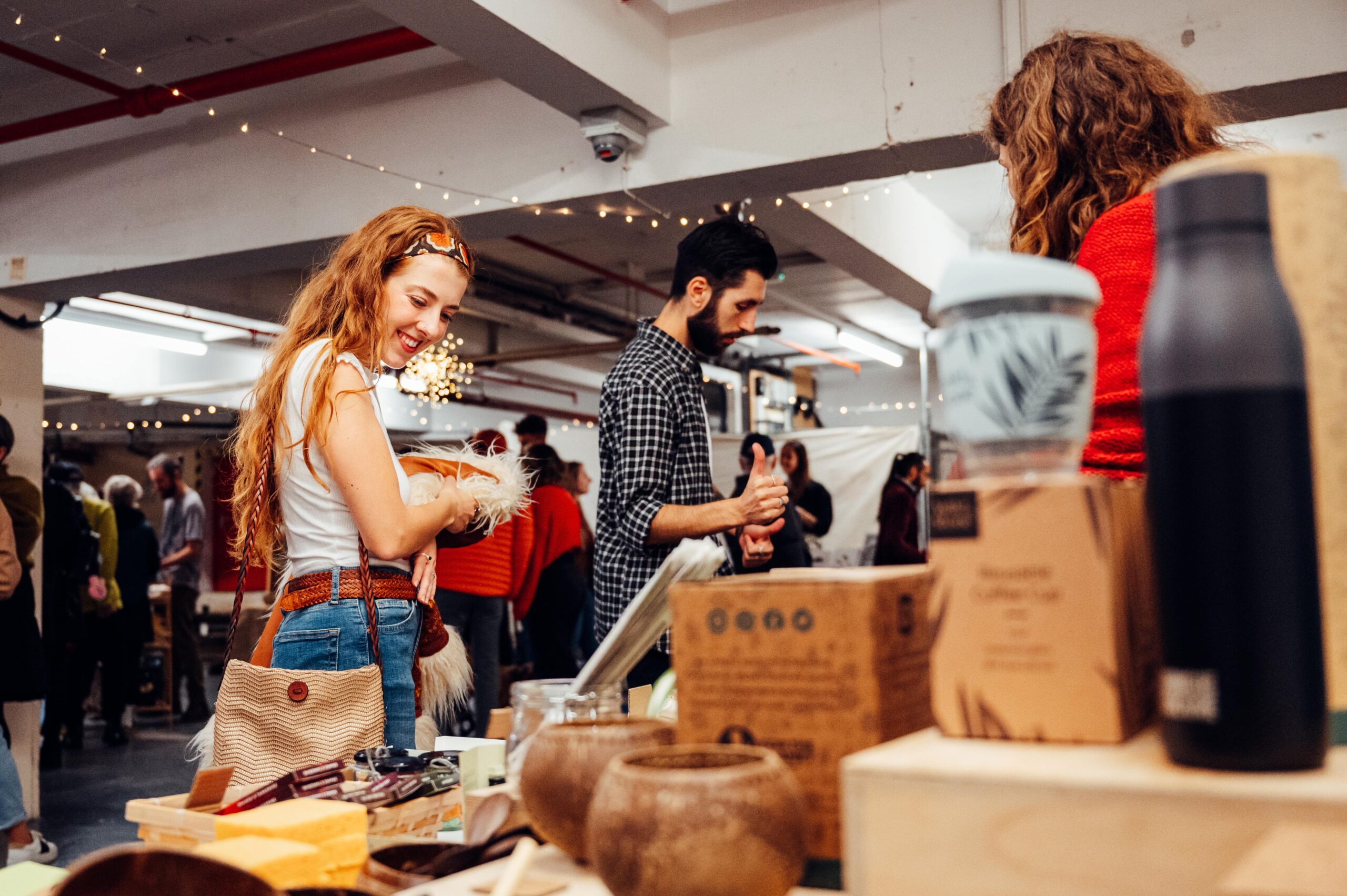 A woman with long hair smiles while browsing items at an indoor market, holding a small, fluffy accessory. The space is decorated with string lights, creating a cozy atmosphere, and other shoppers can be seen in the background exploring various stalls and products.