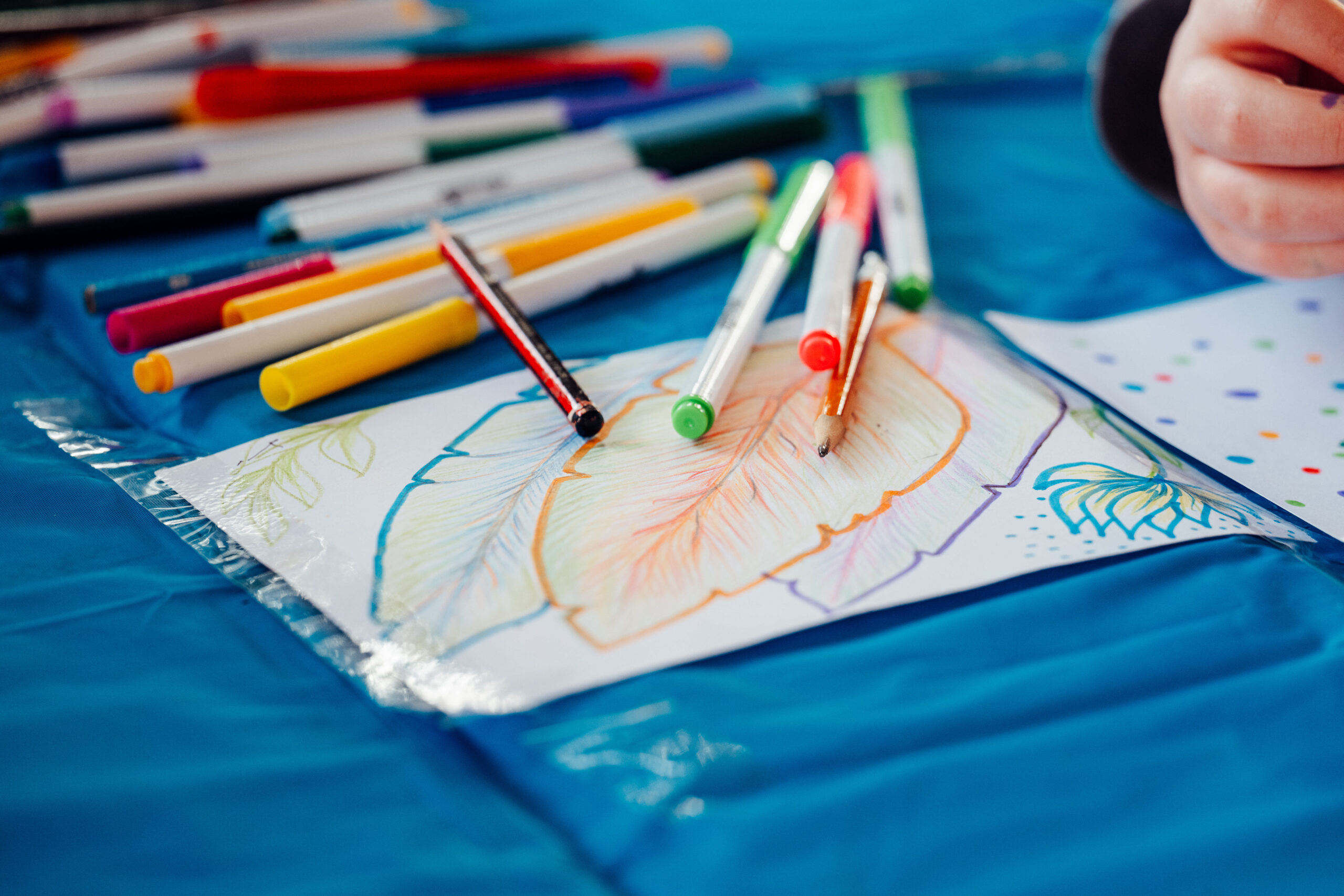 This image shows a creative scene where multiple colored markers and pens are scattered around a table, with a partially completed drawing of a leaf in the foreground. The artwork features vibrant shades of orange, yellow, and blue, and the hand of the artist can be seen in the corner, adding further detail to the drawing.