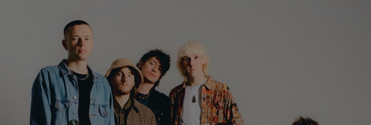 A group of six people with eclectic fashion styles, including denim, patterned shirts, and hats, pose casually in front of a neutral background with a plant in the foreground.