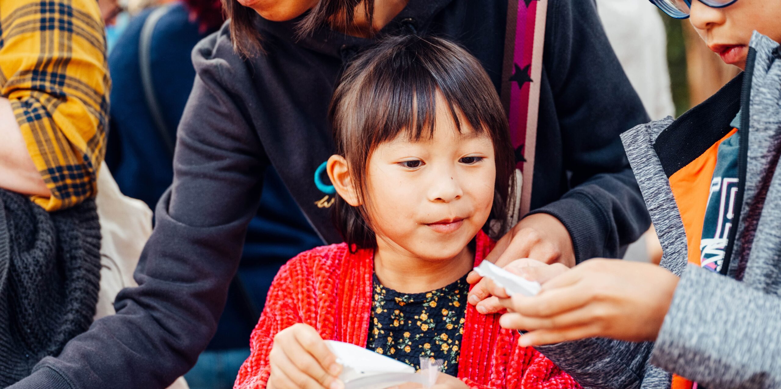 The image features a young girl in a red cardigan and floral dress participating in a craft activity.