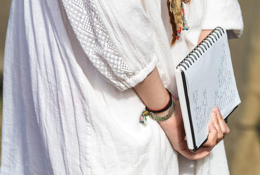 person wearing a white textured dress is holding a spiral notebook against their side.