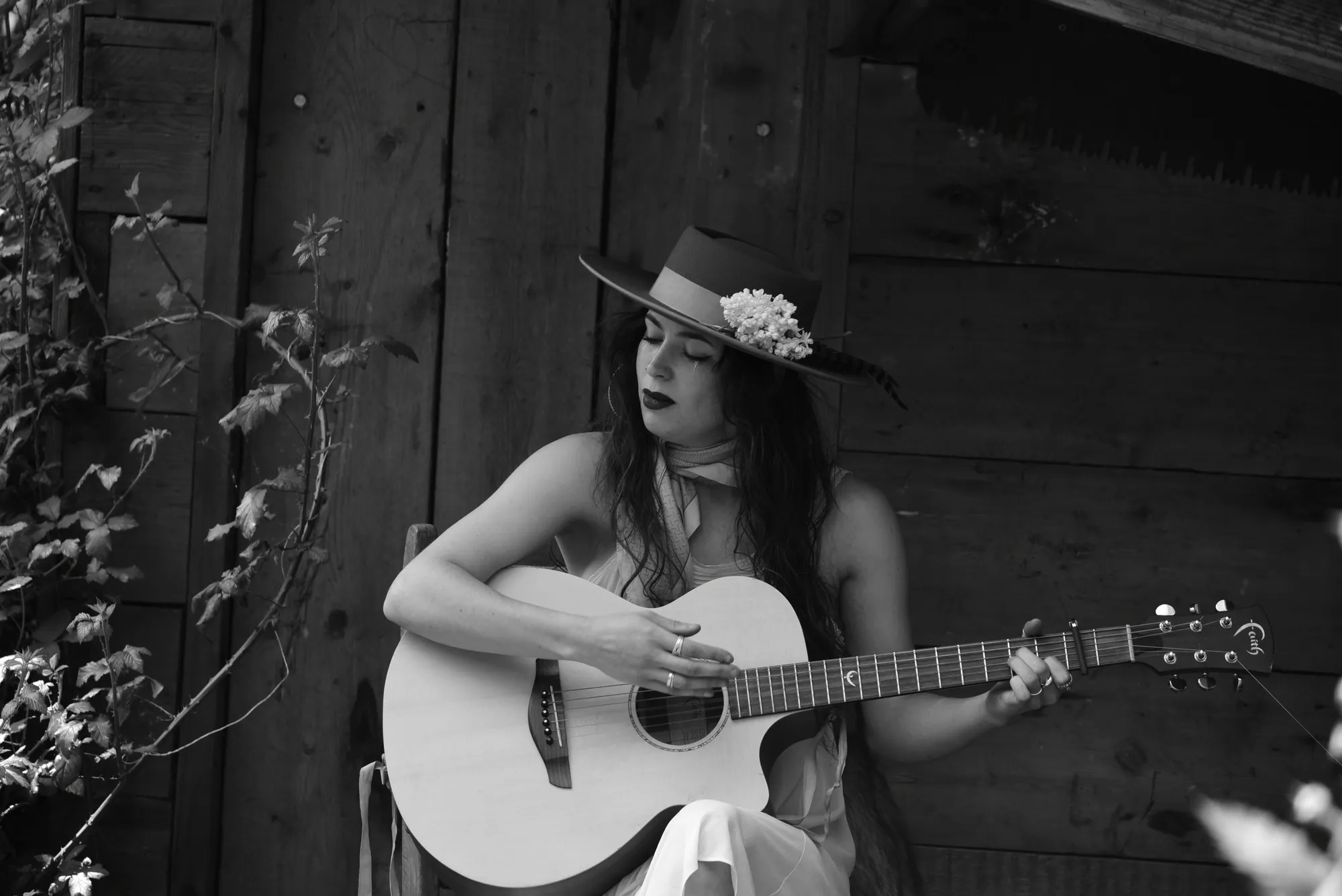 A black-and-white photograph of a woman sitting and playing an acoustic guitar. She is wearing a wide-brimmed hat adorned with flowers, a scarf around her neck, and a light dress.