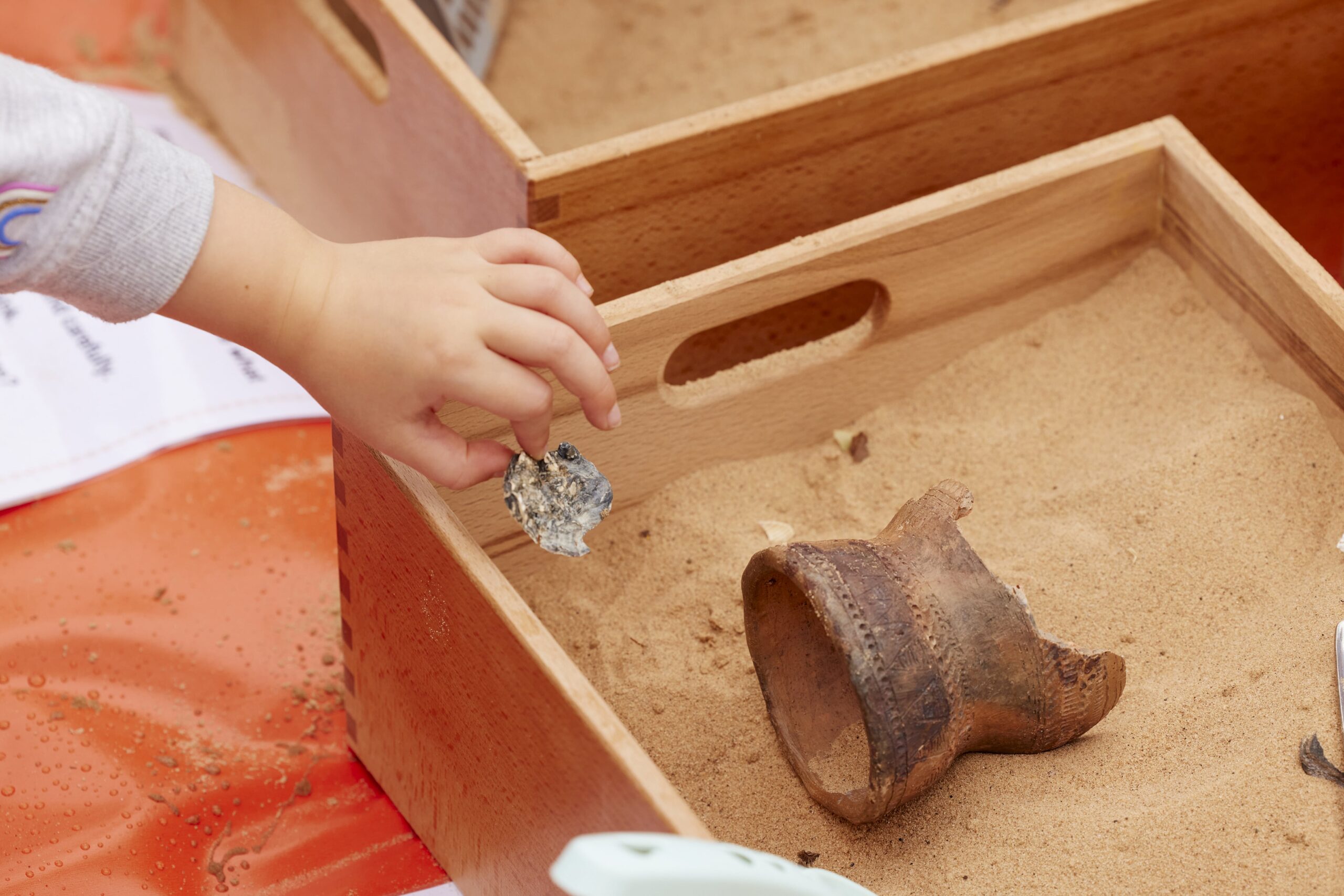 A close-up of a child's hand holding a small artifact over a wooden sandbox.