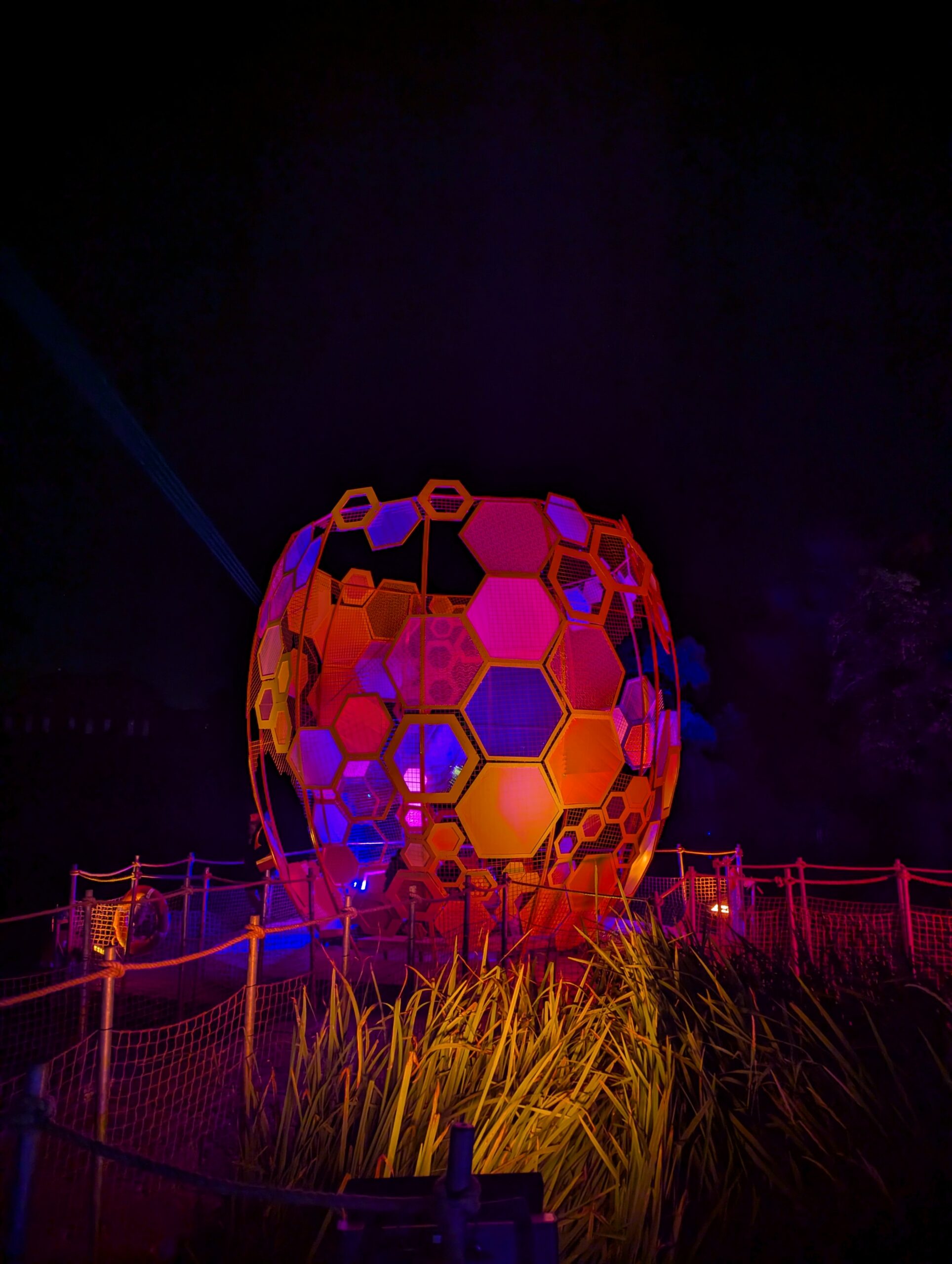 A large, spherical sculpture shaped like a honeycomb composed of hexagonal panels illuminated in vibrant red, orange, and purple hues against a dark night sky.