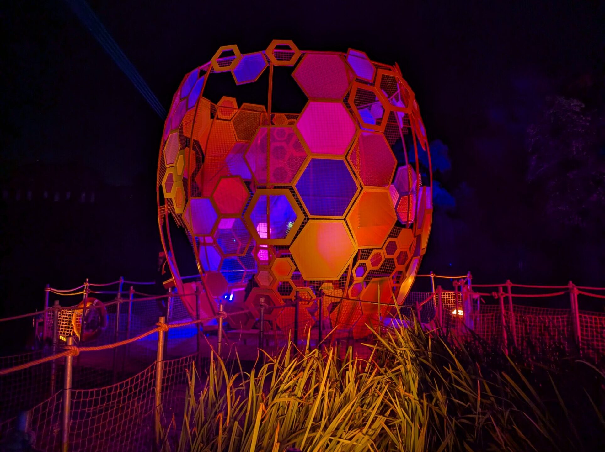 A large, spherical sculpture shaped like a honeycomb composed of hexagonal panels is illuminated in vibrant red, orange, and purple hues against a dark night sky.