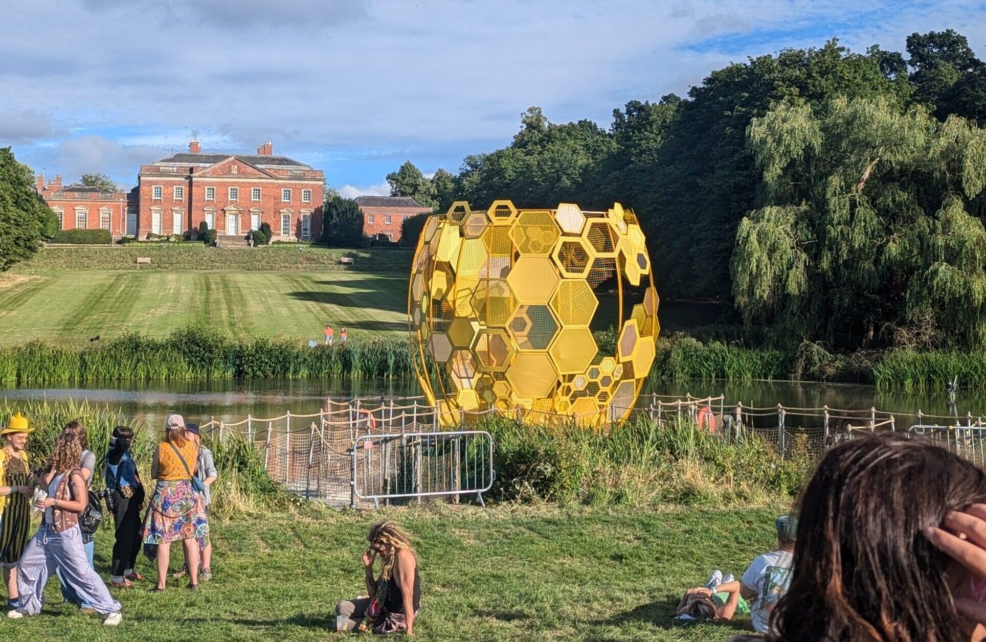 The image shows a large, yellow, spherical sculpture made of hexagonal patterns, resembling a beehive, situated near a pond in a grassy area. In the background, a stately red-brick manor house stands on a hill, surrounded by trees.