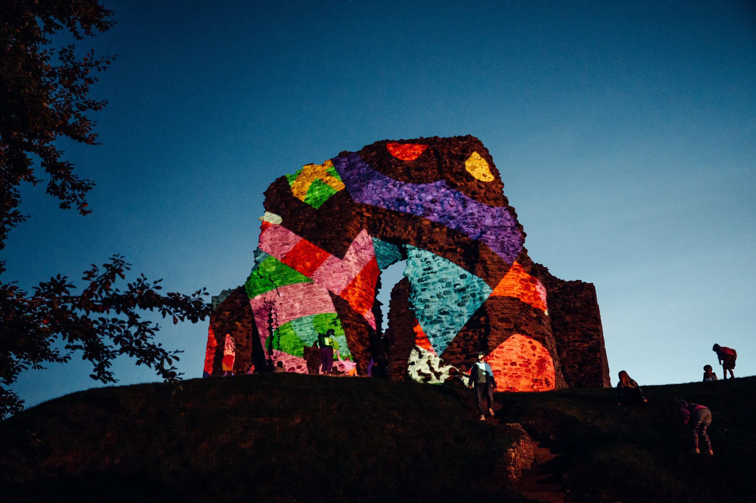 The image shows an ancient stone ruin illuminated with vibrant, abstract geometric patterns in a mix of colors including purple, green, red, yellow, and blue. It appears to be taken at dusk or nighttime, with the sky a deep blue.