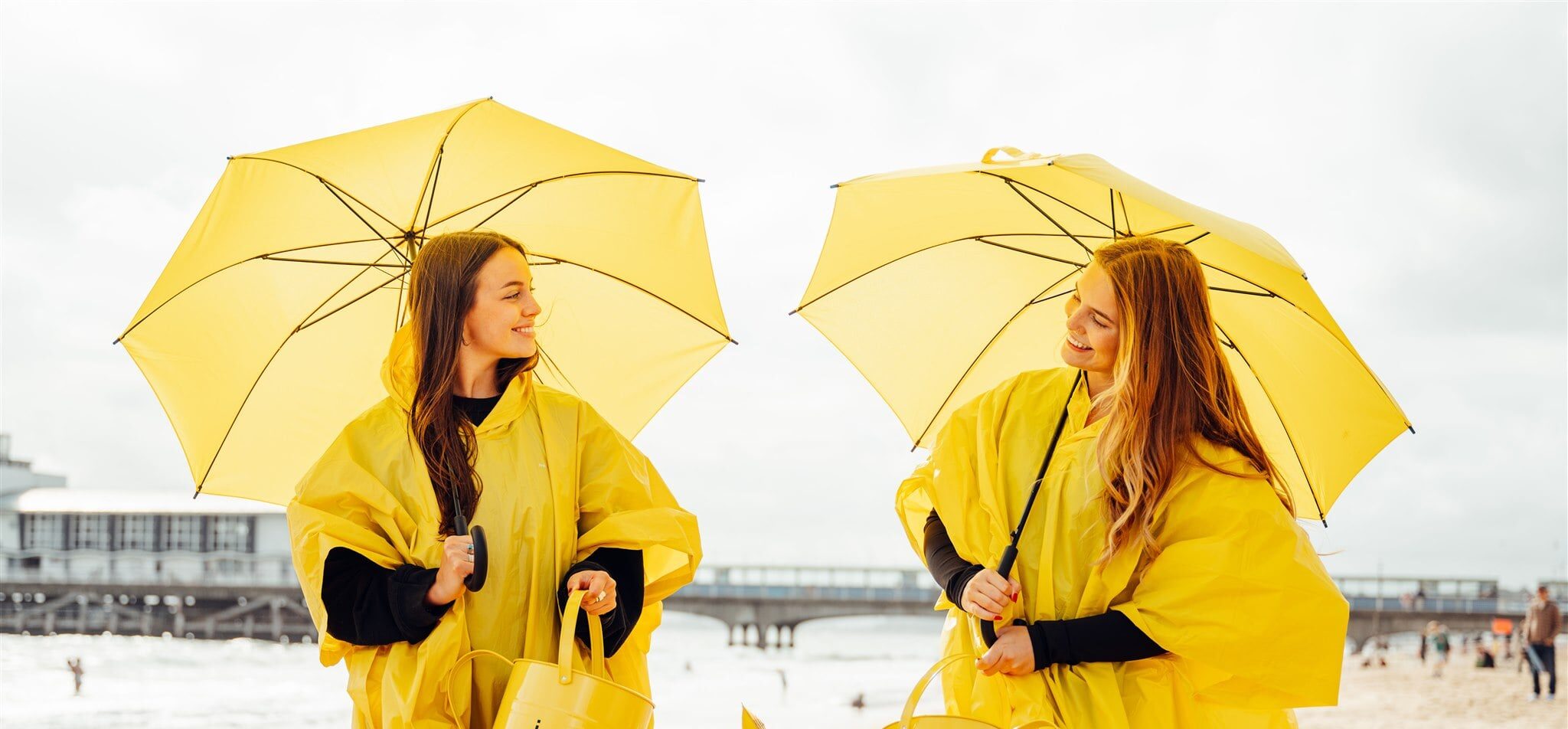 Two girls walk along the beach wearing bright yellow rain coats, holding bright yellow umbrellas and bright yellow watering cans which have the words 'rain trail' written on them