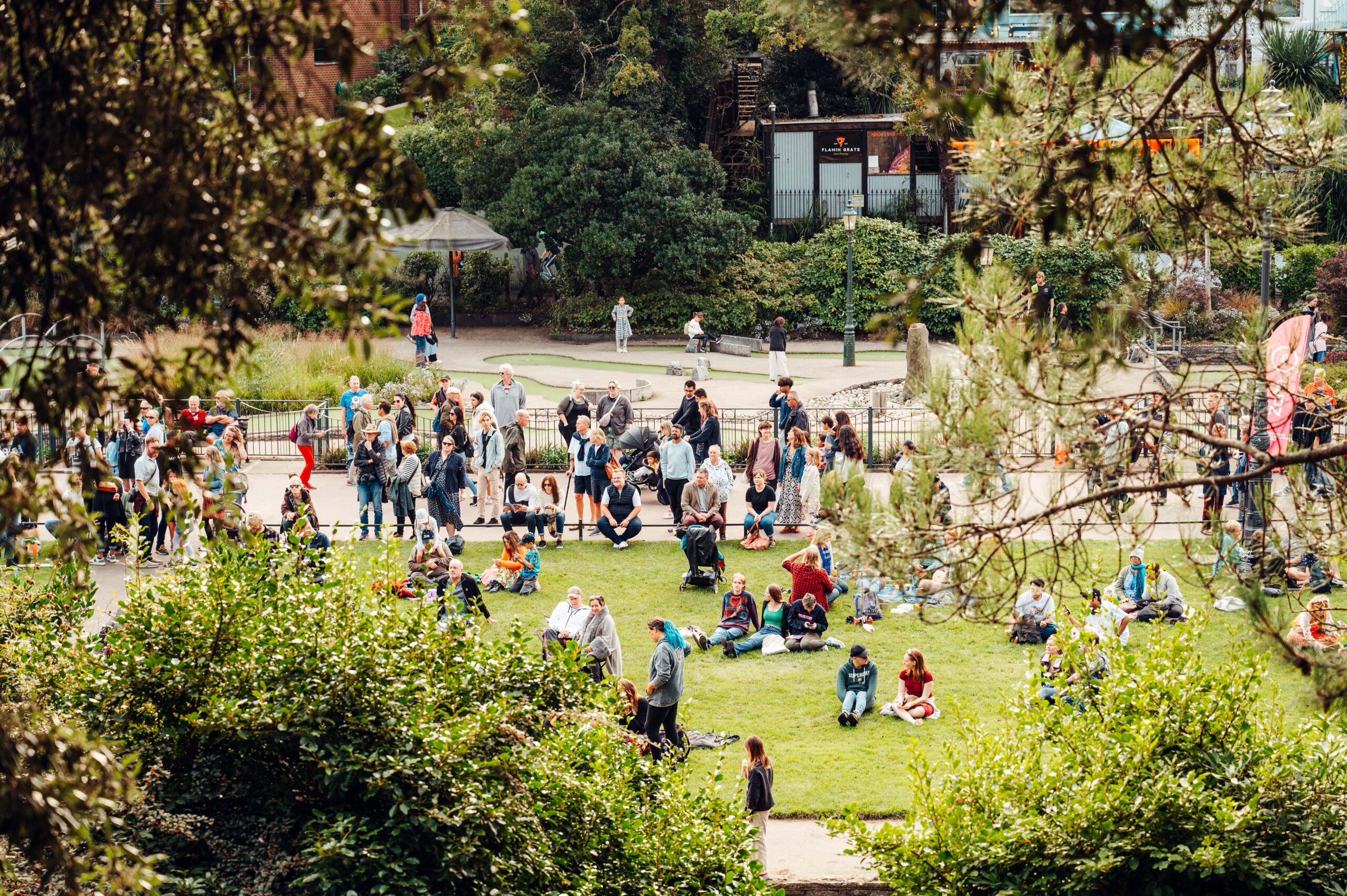 A wide shot of a crowd gathered in a park during the Bournemouth Arts by the Sea Festival. The scene is framed by greenery, with people standing, sitting, and relaxing on the grass. In the background, there are trees, shrubs, and festival signage. Some individuals are engaged in conversation, while others sit on the ground in small groups, enjoying the festival atmosphere.