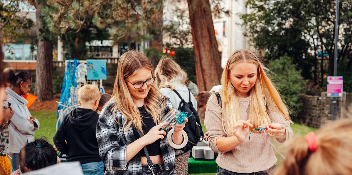 Two women engage in a craft activity outdoors, surrounded by colorful materials on a table. One woman with glasses holds a clear container filled with blue craft materials, while the other inspects some colored ribbons.