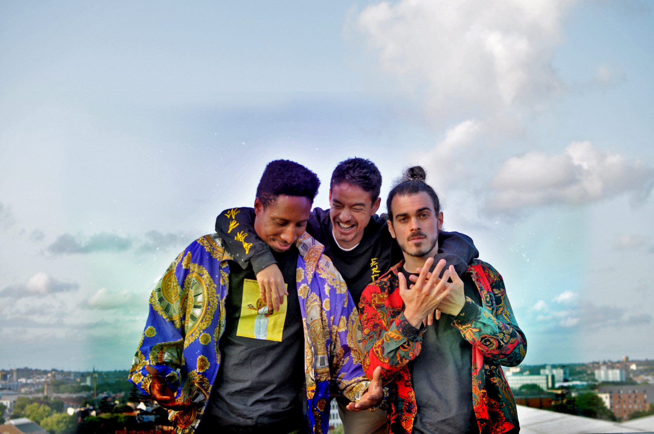 Three men pose together outdoors, smiling and laughing. One has his arm around another’s shoulder, while the third displays a hand gesture. They wear brightly patterned jackets and black T-shirts, with a clear sky in the background.