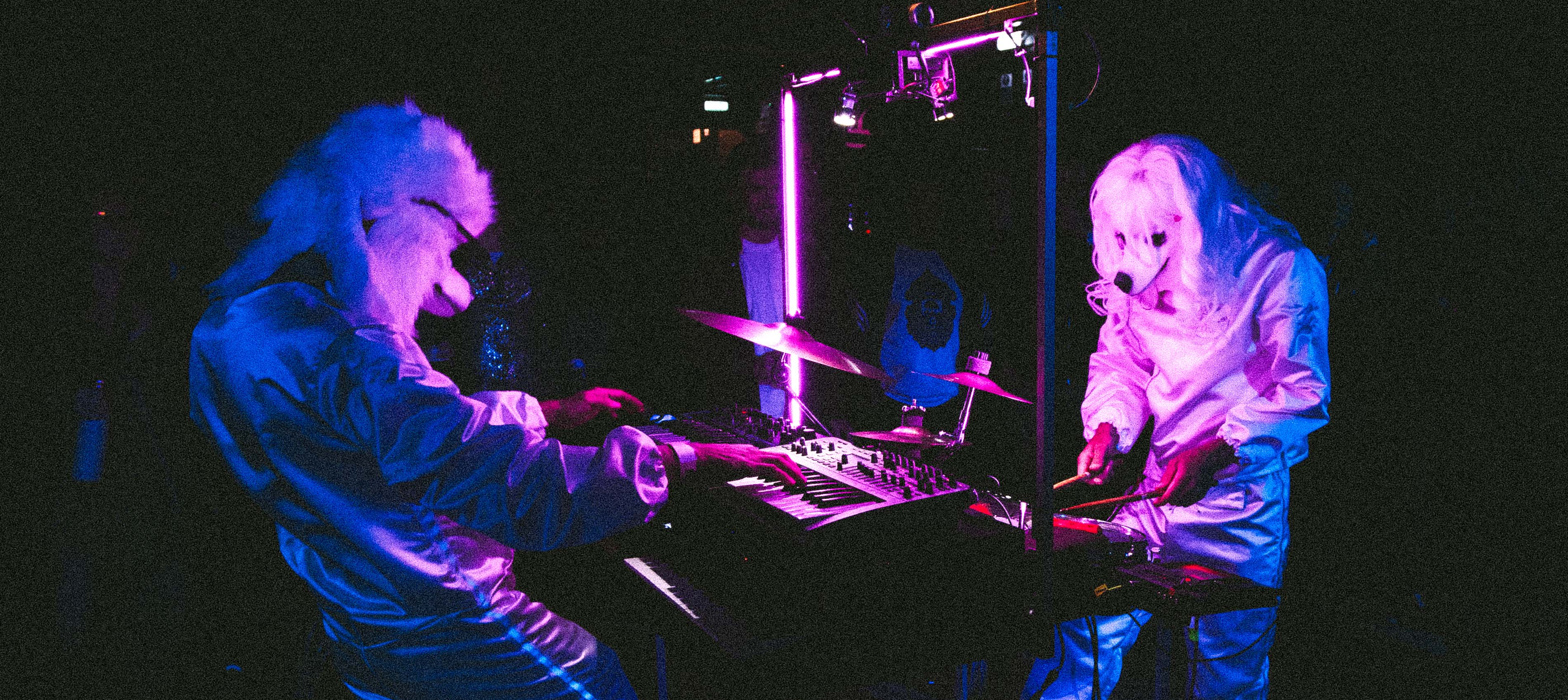 Two performers in white dog costumes play electronic instruments under neon lights.