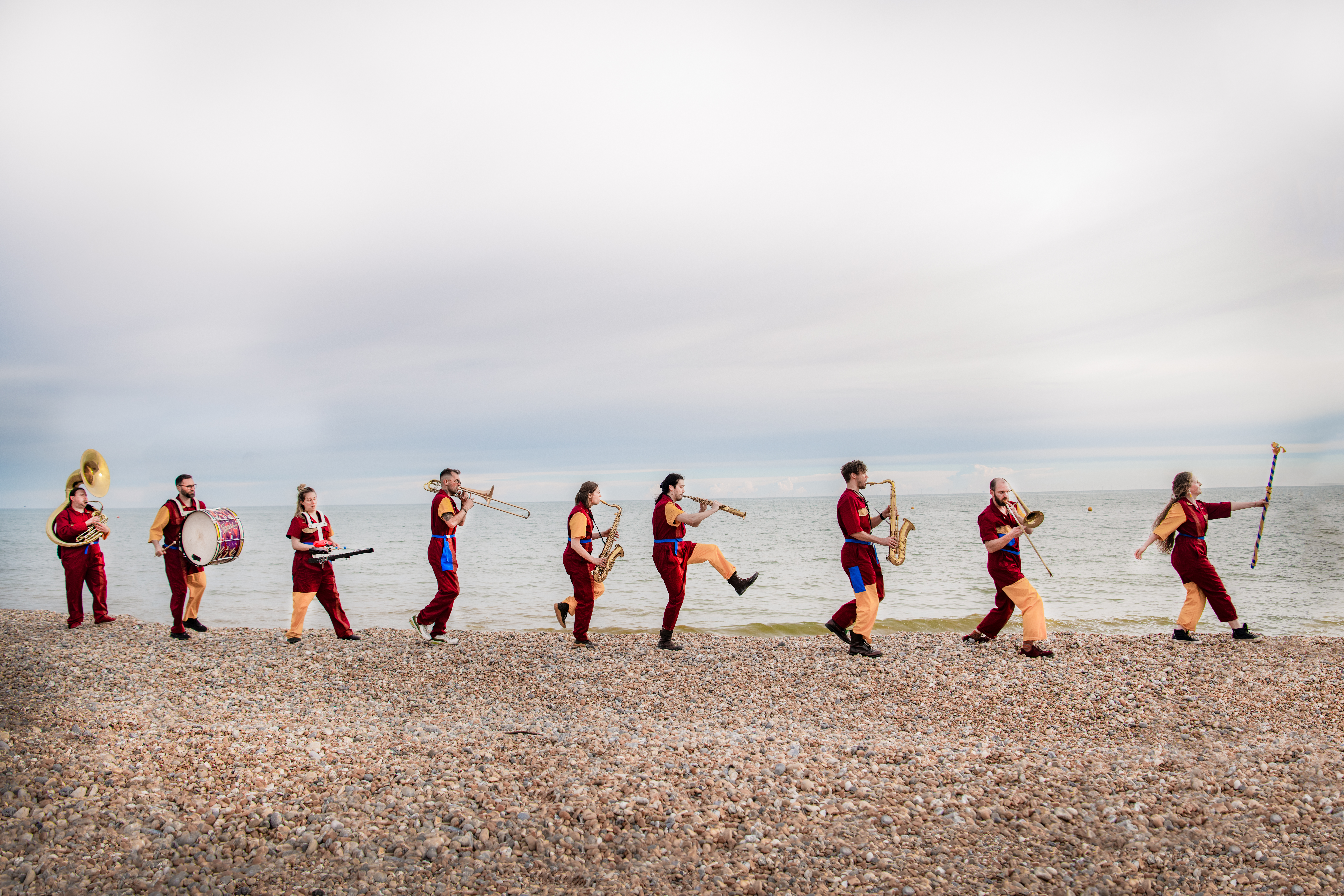 Brass band in colorful uniforms marching along a pebbled beach by the sea.