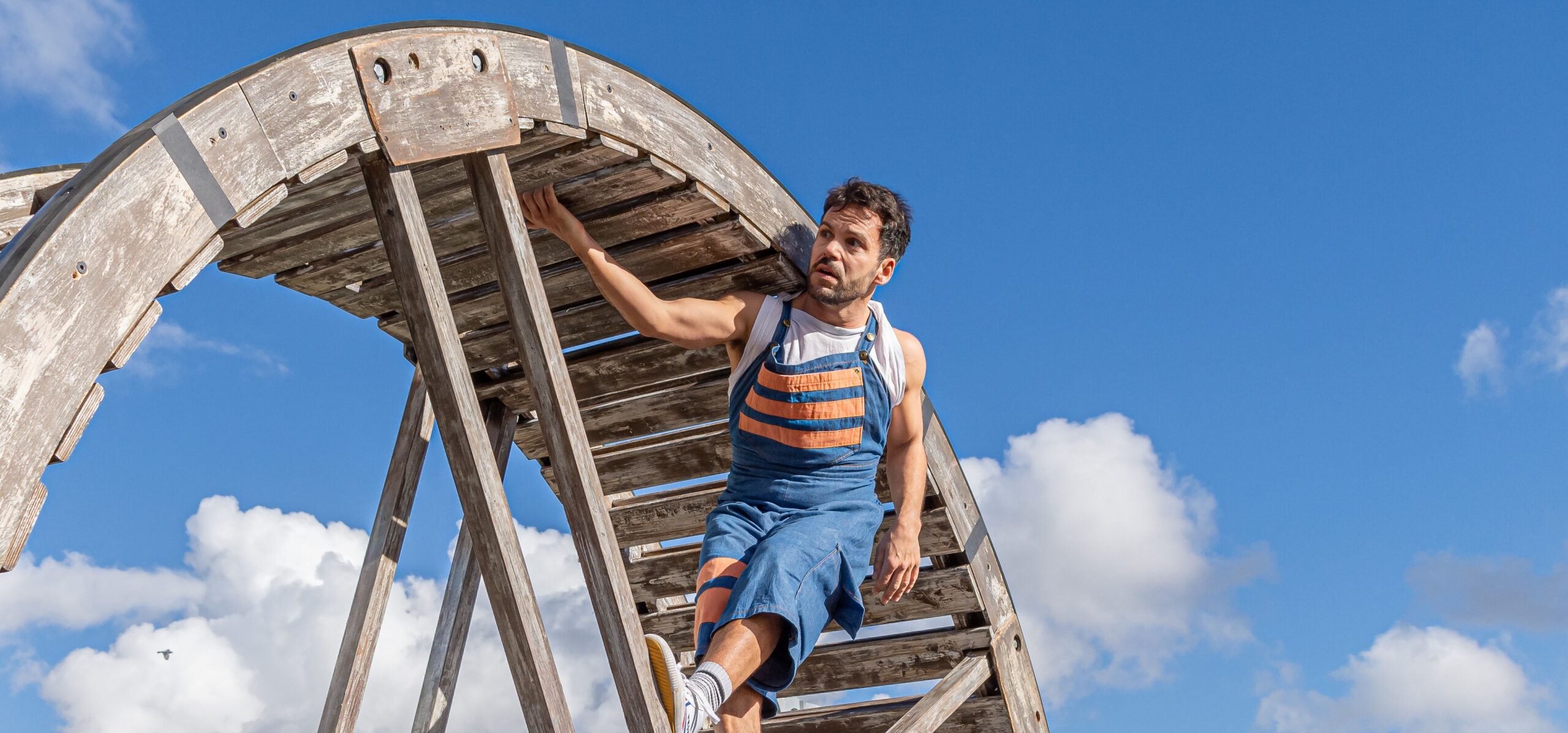 A male performer in blue and orange overalls hangs from the inside of a large wooden wheel structure against a bright blue sky with scattered clouds. He grips an overhead beam while balancing on the wheel’s frame, appearing mid-performance.