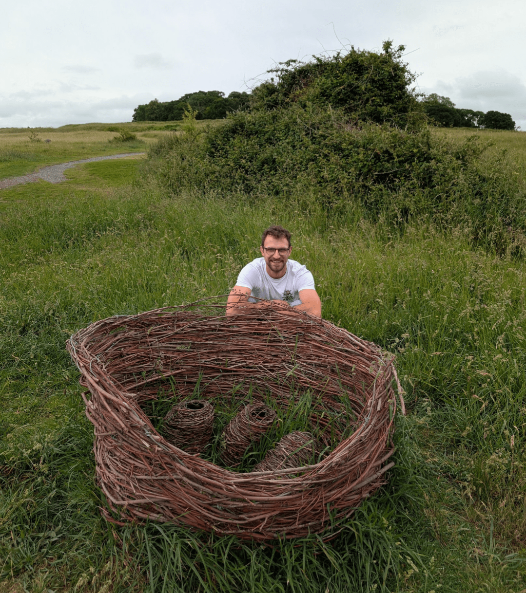 Man smiles behind a large woven nest-like sculpture with three forms inside, set in a grassy field with a path and trees beyond.