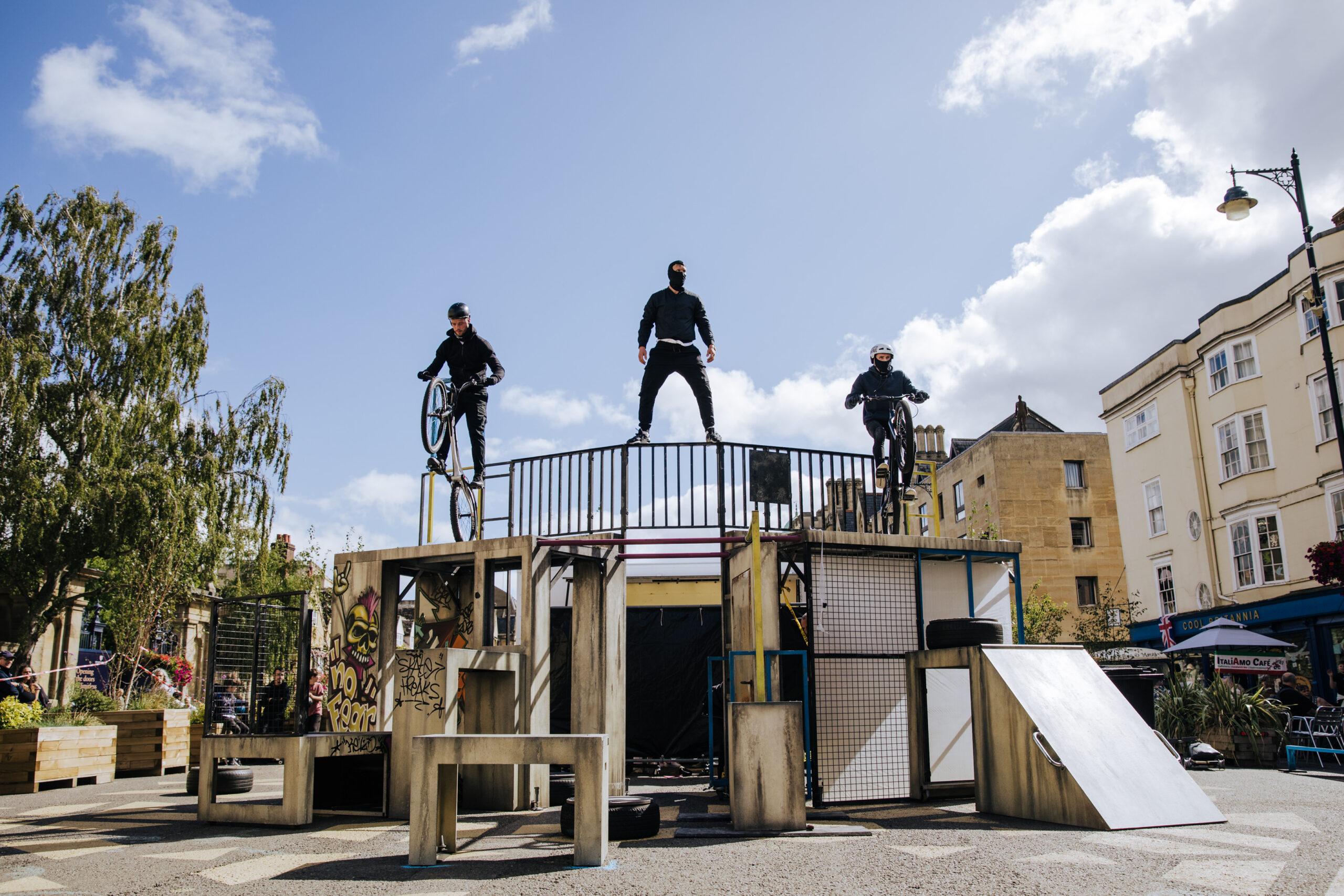 Two stunt cyclists and a performer stand atop an urban stage structure, balancing high above the crowd under a blue sky.