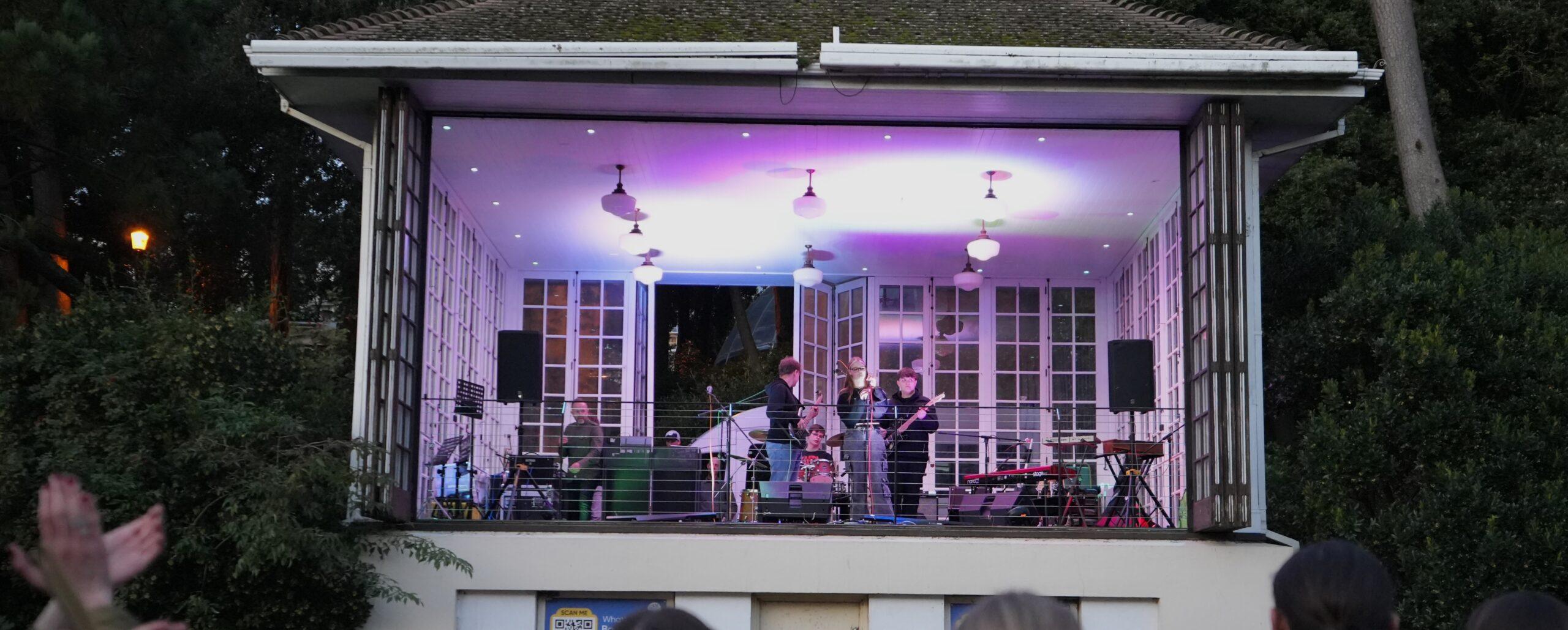 Band performs in a lit bandstand with purple and white lights as audience members watch from outside in the evening.