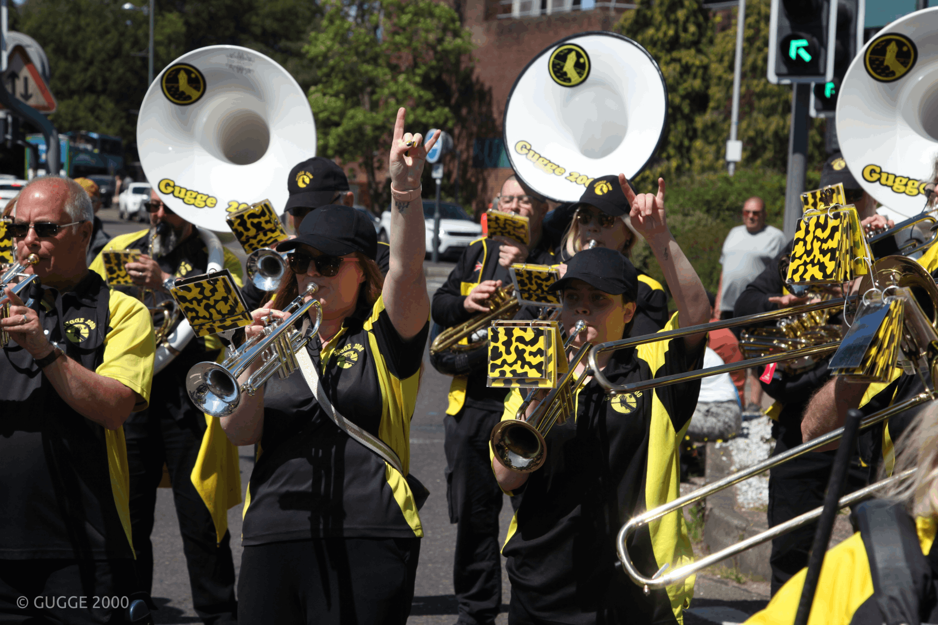 Brass band in black and yellow uniforms plays trumpets, trombones, and sousaphones during an energetic outdoor parade.