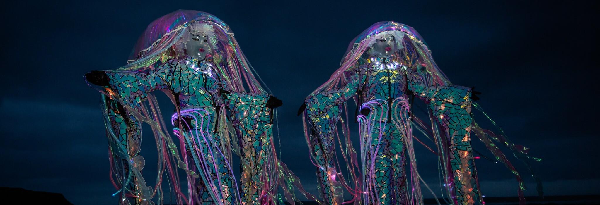 Two glowing jellyfish performers in illuminated costumes with trailing tendrils stand out against the dark night sky.