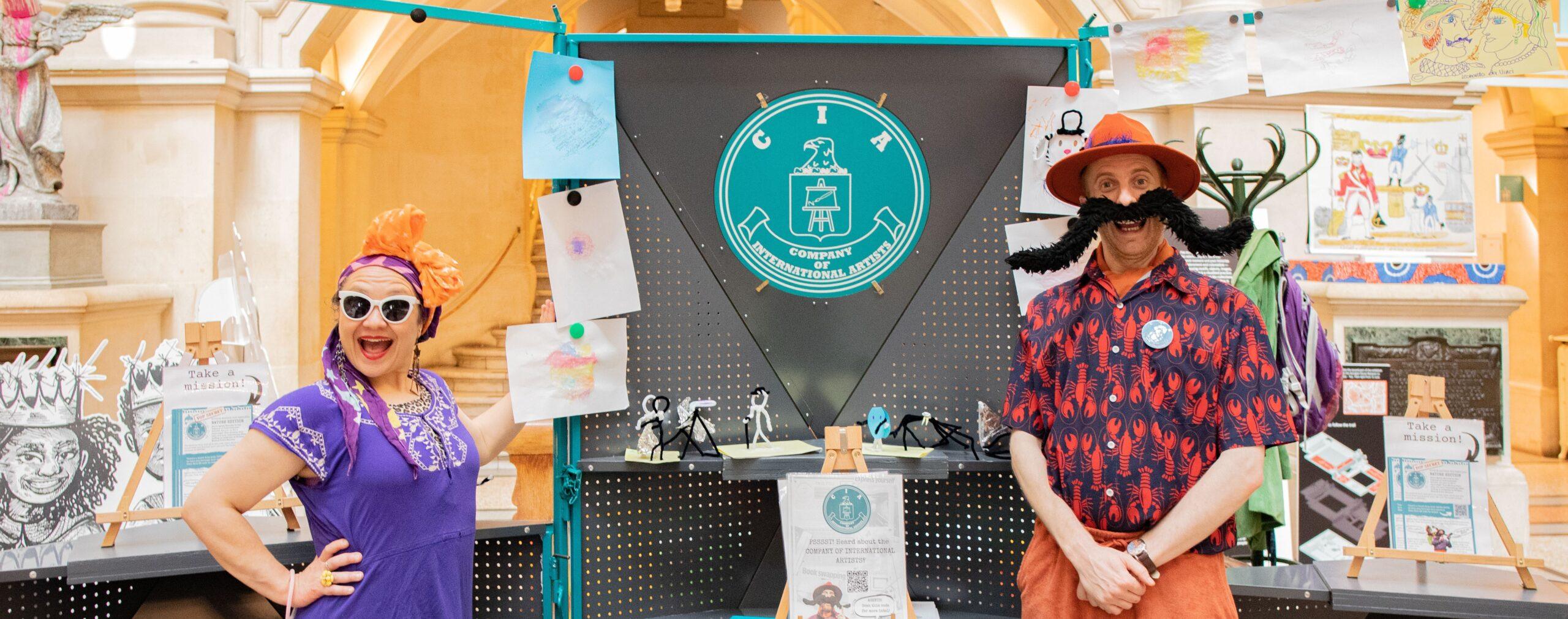 Two performers in bright costumes stand by a “Company of International Artists” display decorated with drawings and signs.