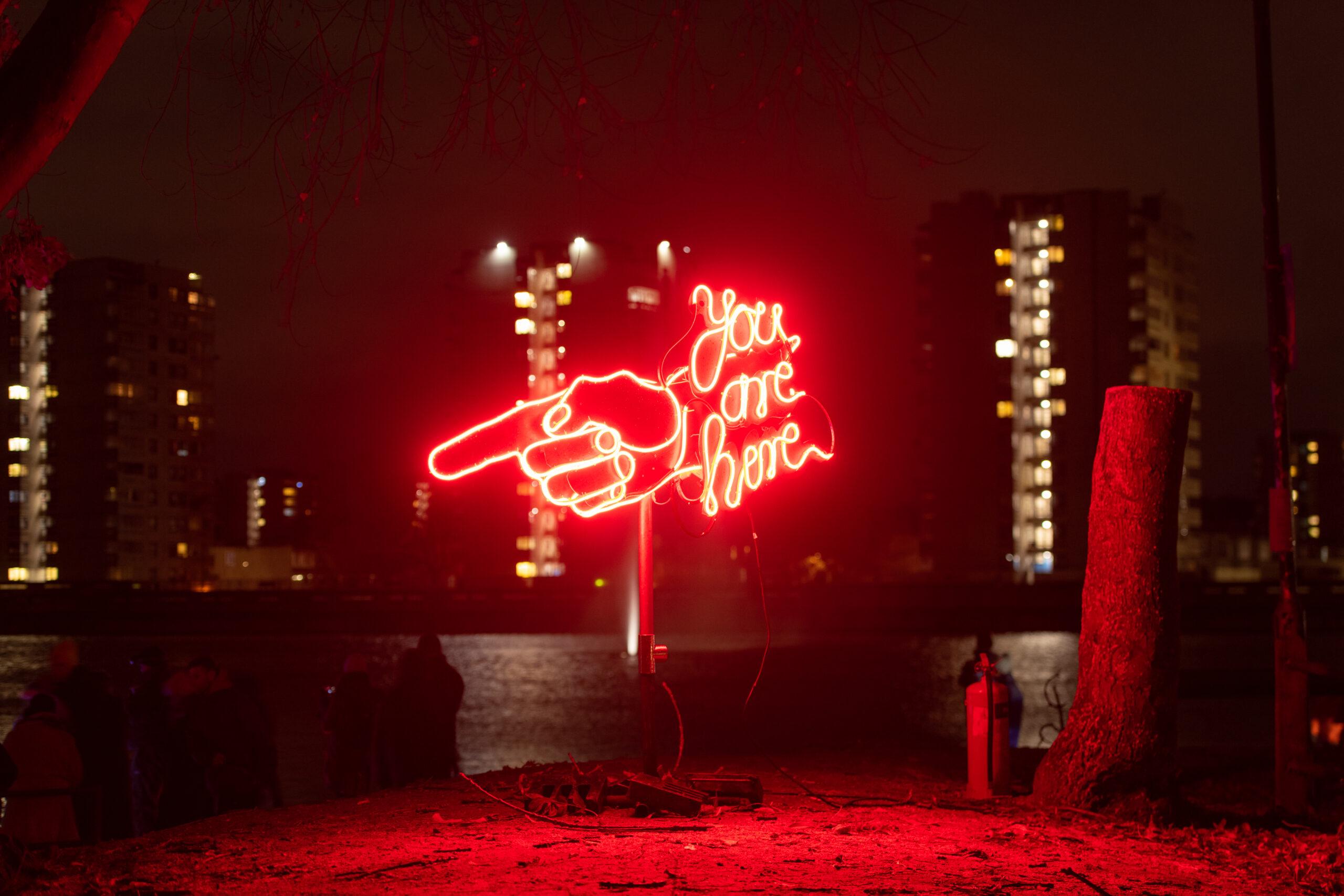 Red neon sign of a pointing hand with the words “You are here,” glowing at night against city tower blocks.
