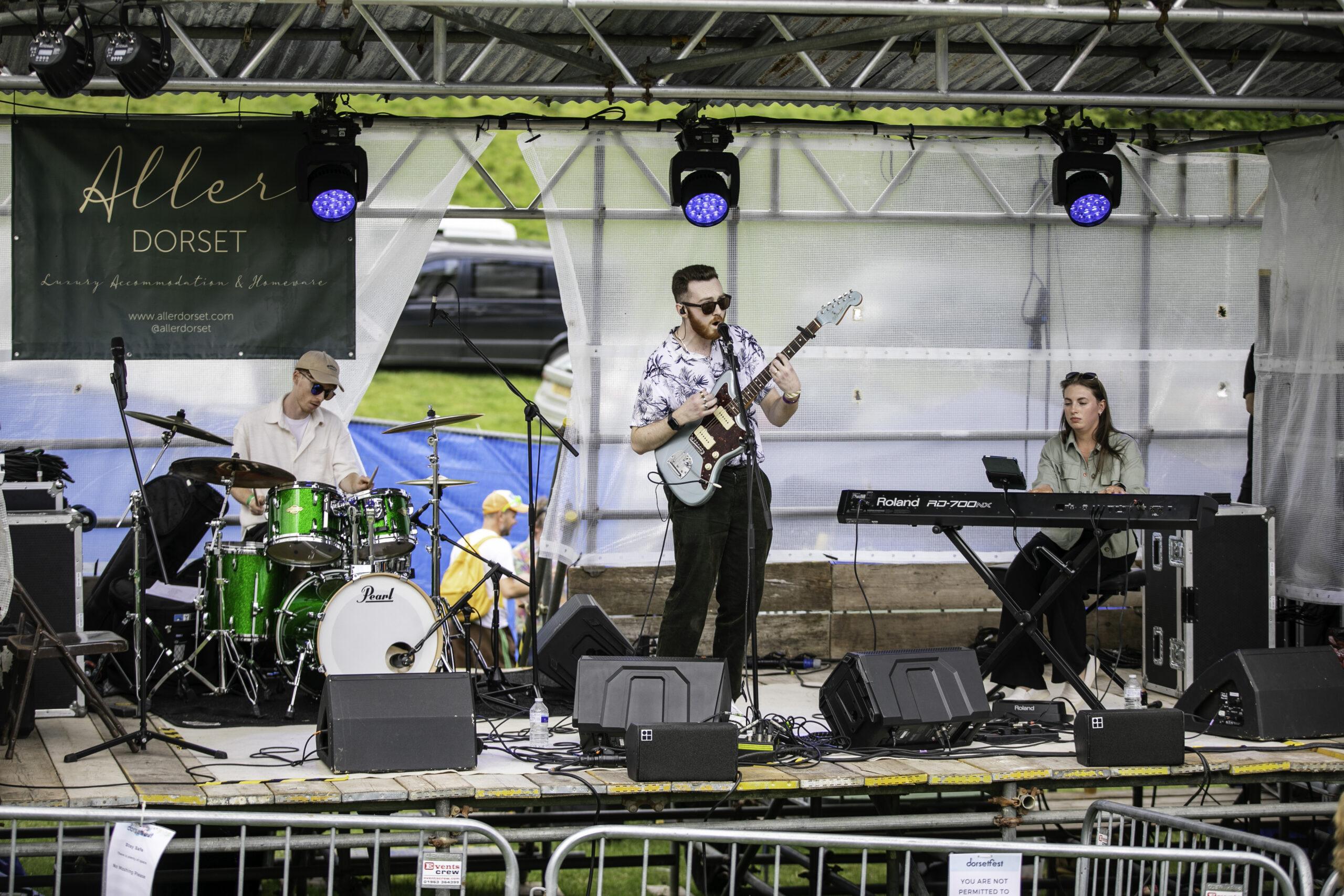Three-piece band performs on an outdoor festival stage with drums, guitar, and keyboard under a banner reading “Aller Dorset.”