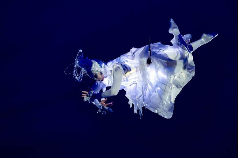 Aerial performer in glowing white costume and headdress floats suspended against a dark blue background, arms reaching outward.